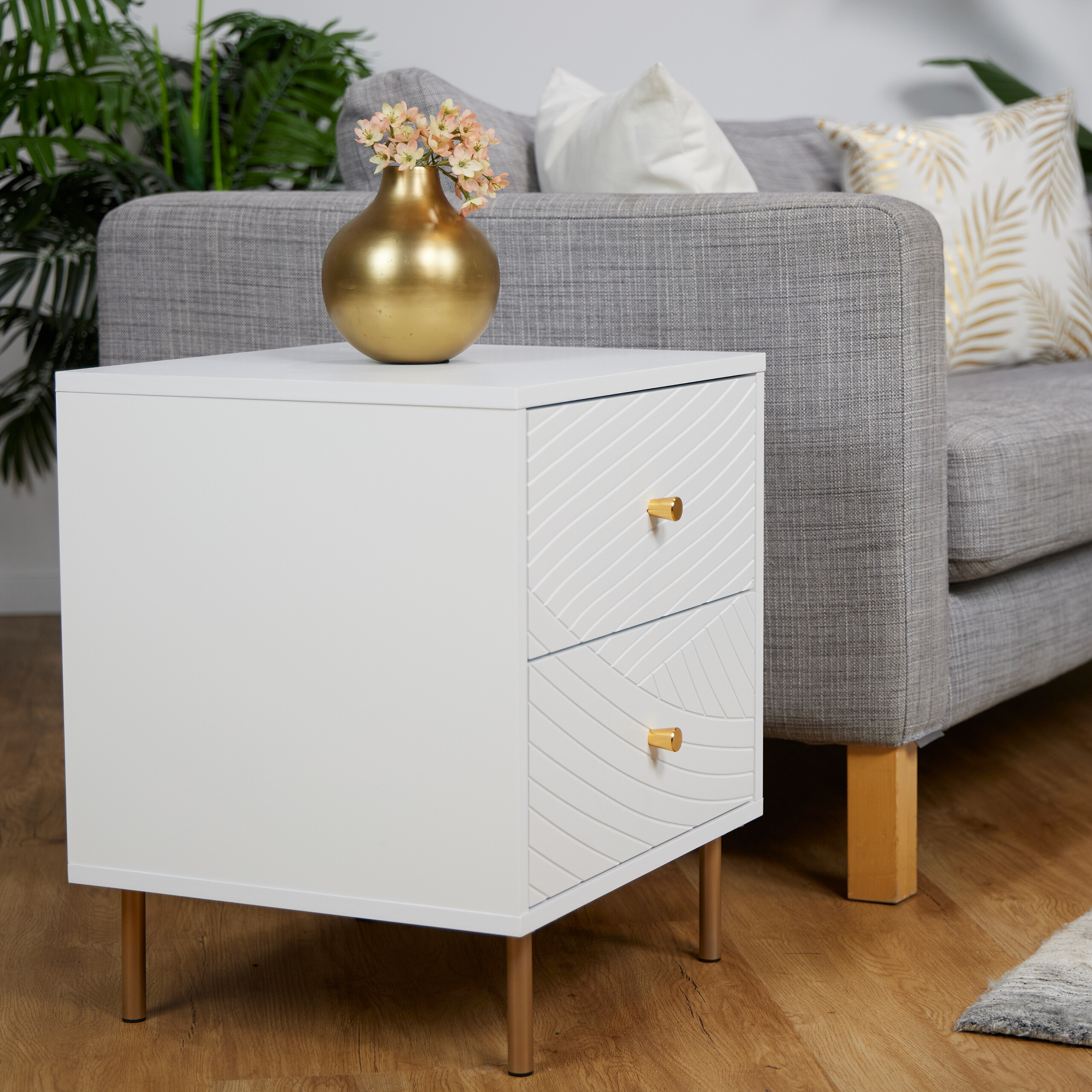 Ein weißer Nachttisch mit goldenen Beinen und einer goldenen Vase neben einem grauen Sofa.