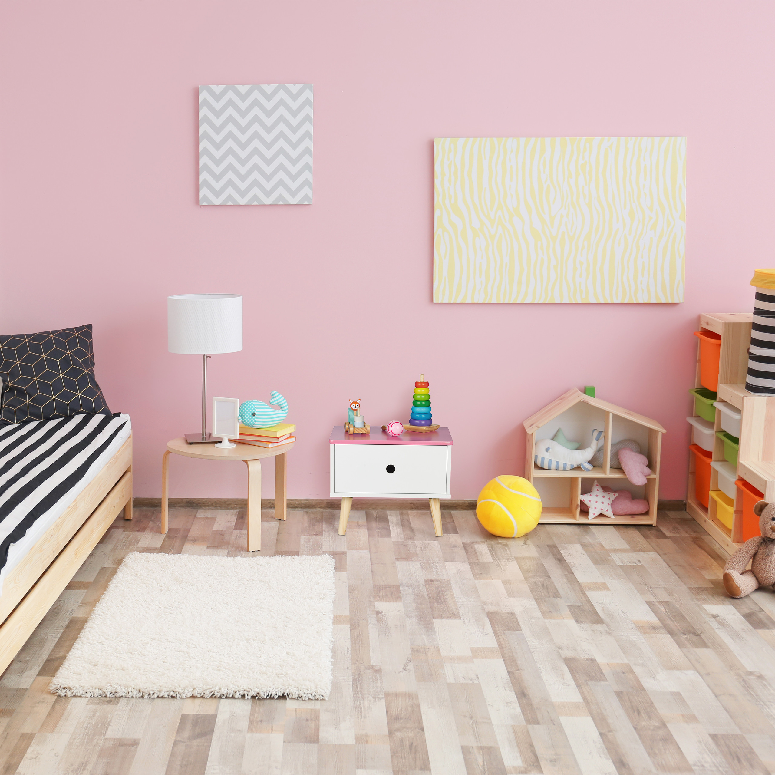 Ein rosafarbenes Kinderzimmer mit Bett, Nachttisch, Lampe, Spielzeug und einem Holzboden.