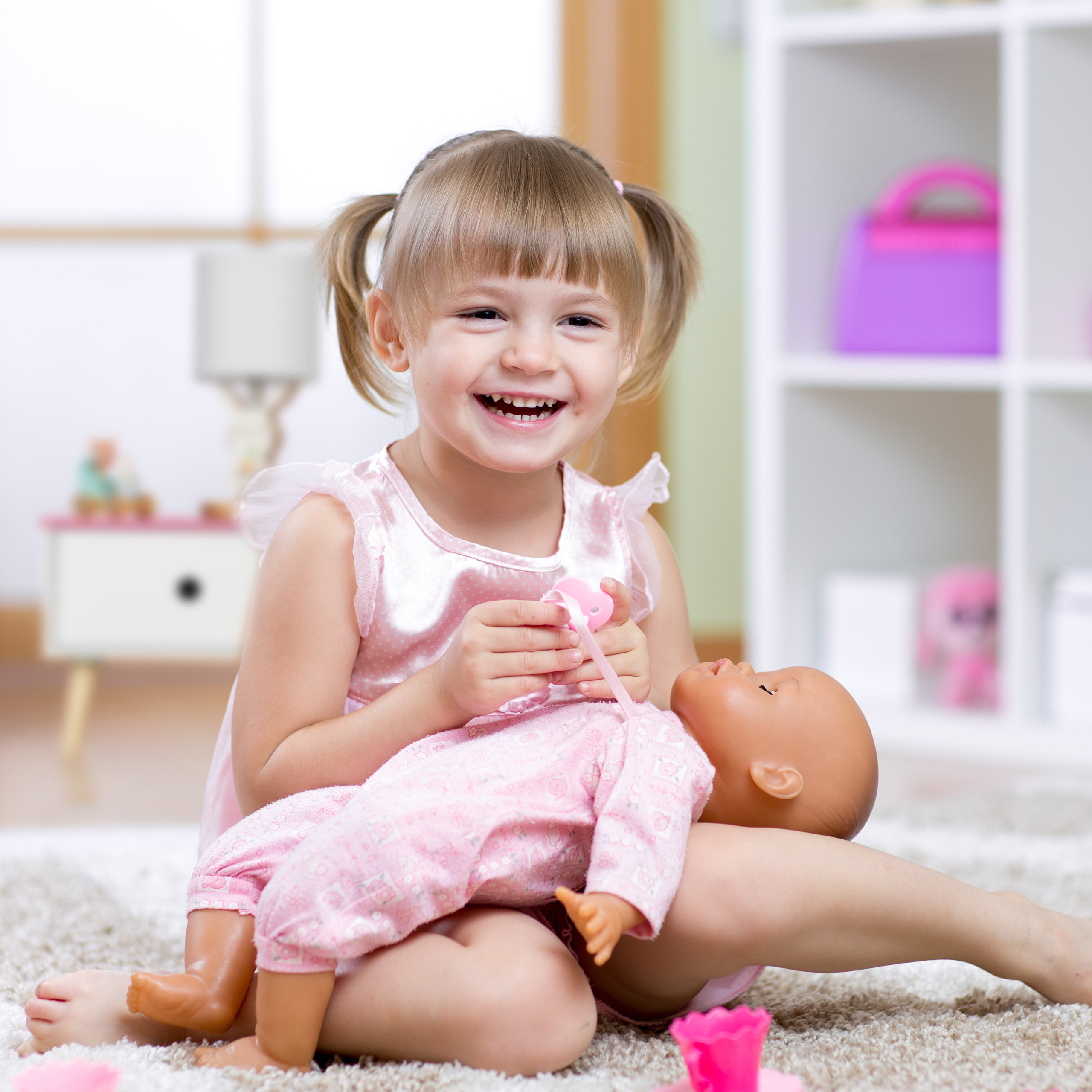 Ein lächelndes Mädchen im pinken Kleid spielt mit einer Puppe auf einem Teppich in einem hellen Zimmer.
