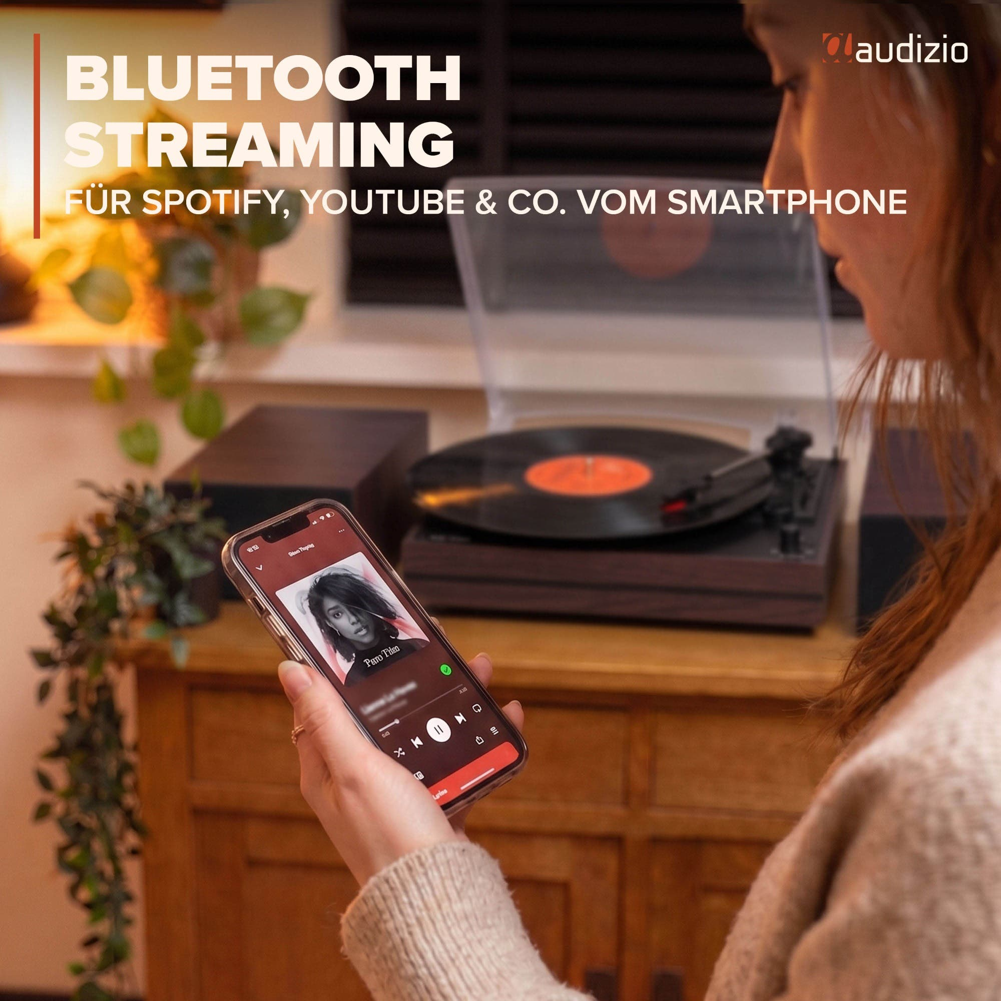 Una mujer sostiene un móvil con app de música, con un tocadiscos y altavoces sobre un mueble de madera al fondo.