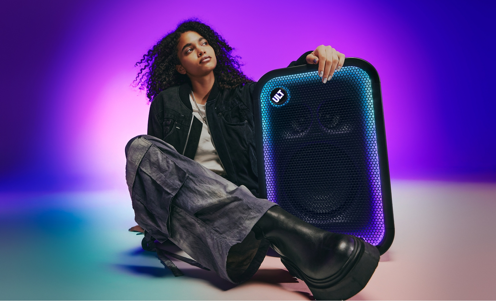 Una mujer con cabello rizado se sienta junto a un gran altavoz con luces azules brillantes sobre fondo morado.