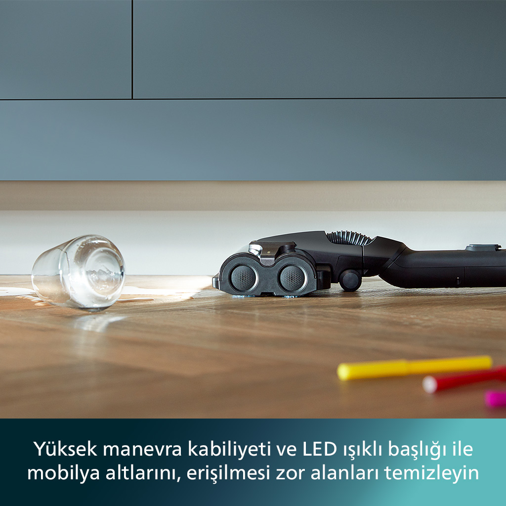 Siyah bir süpürge başlığı, dökülmüş bir bardak su ve renkli kalemlerin yakınında tahta zemini temizliyor.