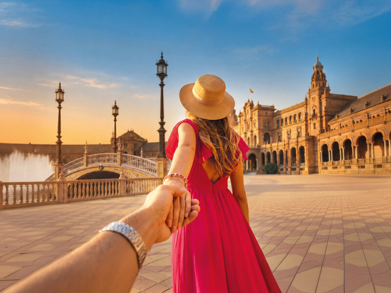 Una mujer con vestido rojo guía a su pareja de la mano frente a una pintoresca arquitectura histórica.