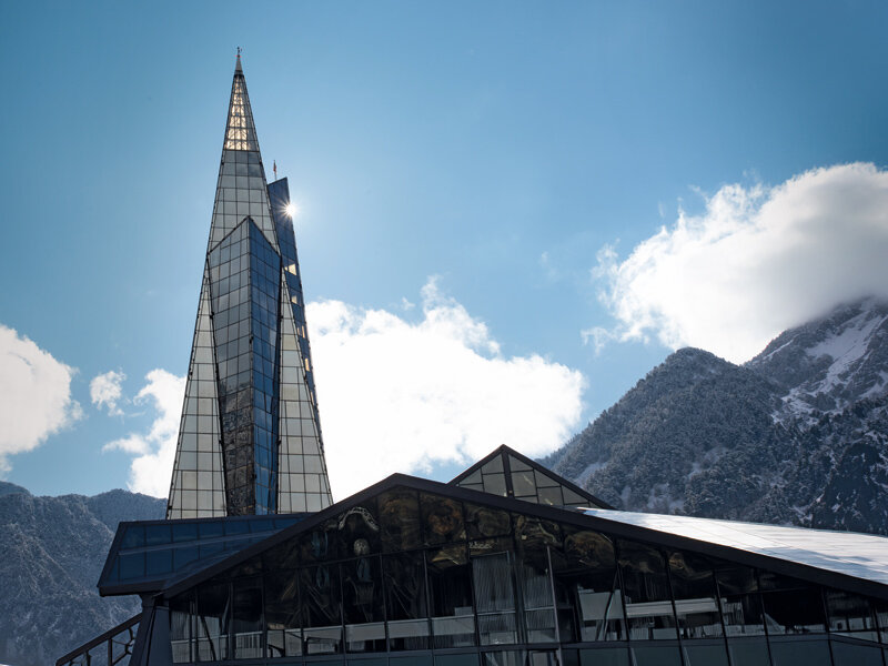Iglesia moderna con torre de vidrio puntiaguda frente a montañas nevadas y cielo azul.