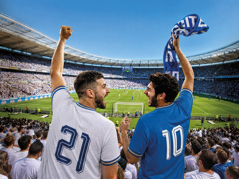 Dos aficionados al fútbol con camisetas celebran con los brazos levantados en un estadio lleno.