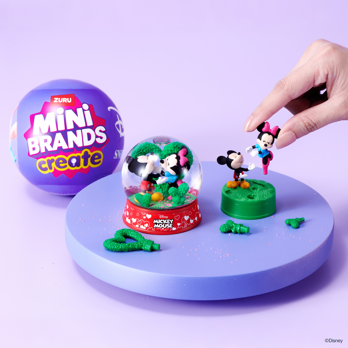 Una mano coloca figuras de Mickey y Minnie Mouse cerca de una bola de nieve en una superficie morada con una cápsula.