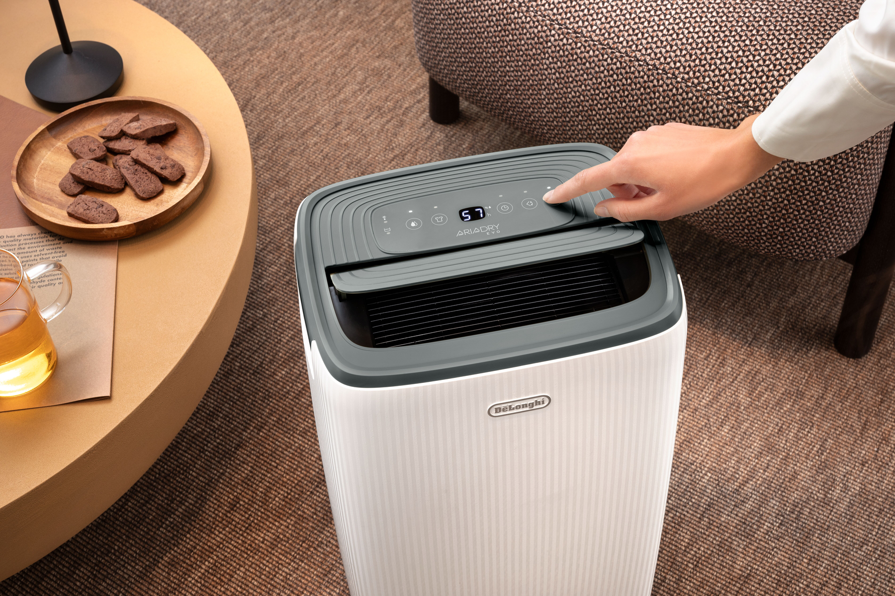 Mano operando la pantalla digital de un deshumidificador blanco DeLonghi en alfombra marrón cerca de mesa con galletas.