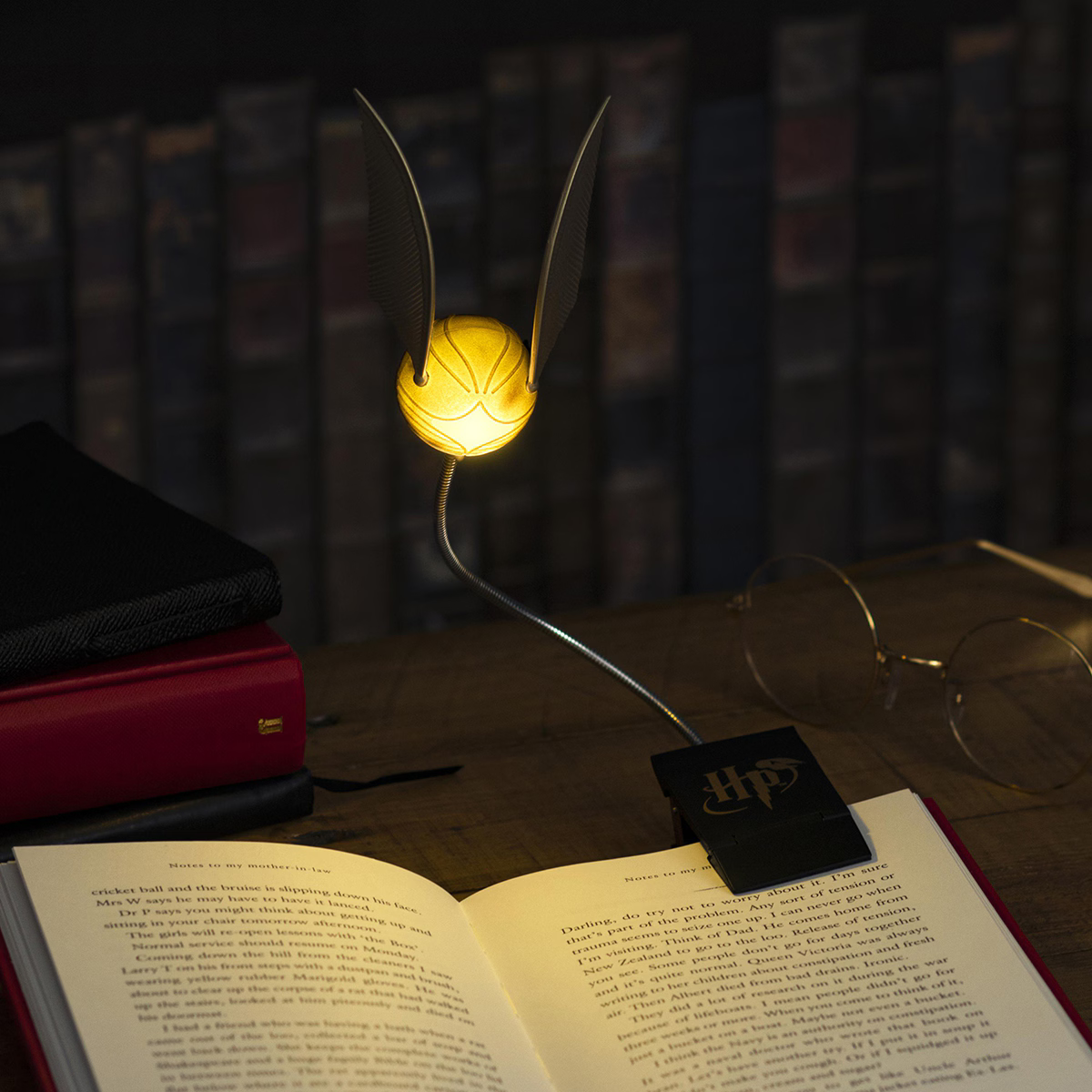 Una luz de lectura de la Snitch Dorada brilla sobre un libro abierto, con gafas redondas y libros al fondo.