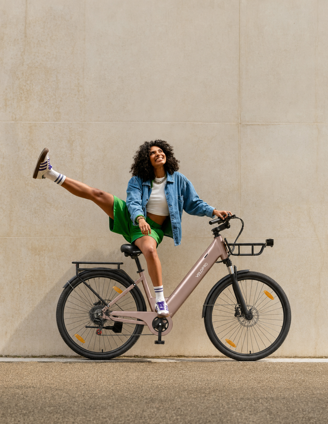 Eine Frau in Jeansjacke und grünen Shorts sitzt auf einem rosefarbenen E-Bike und hebt ein Bein.