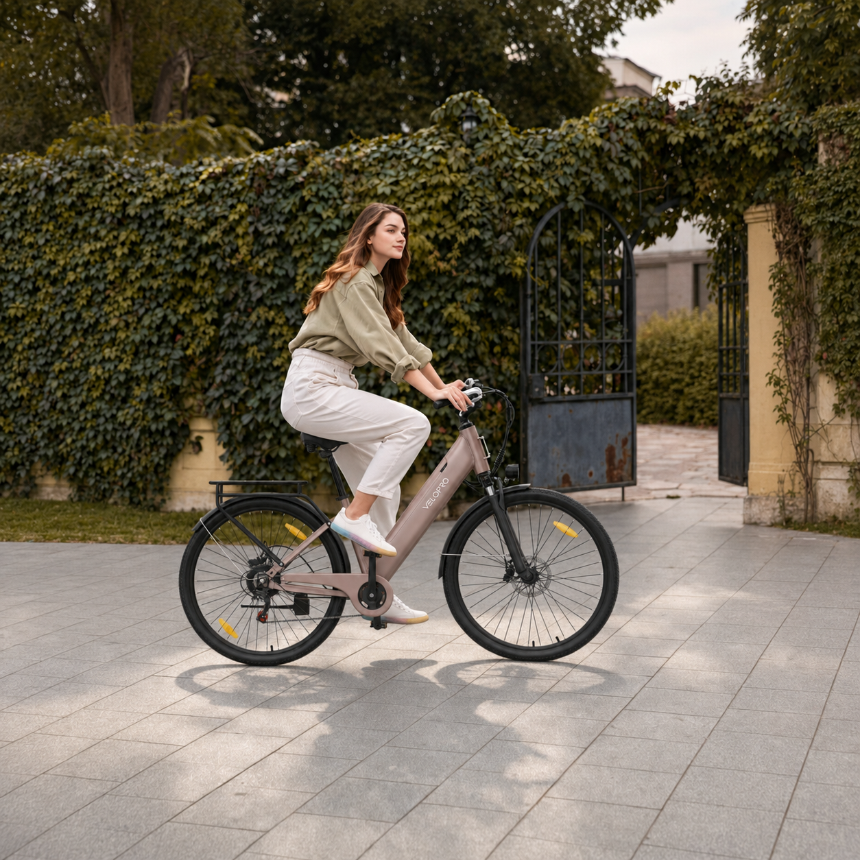 Eine Frau in heller Kleidung fährt auf einem modernen Elektrofahrrad auf einem Steinweg vor einer grünen Hecke.