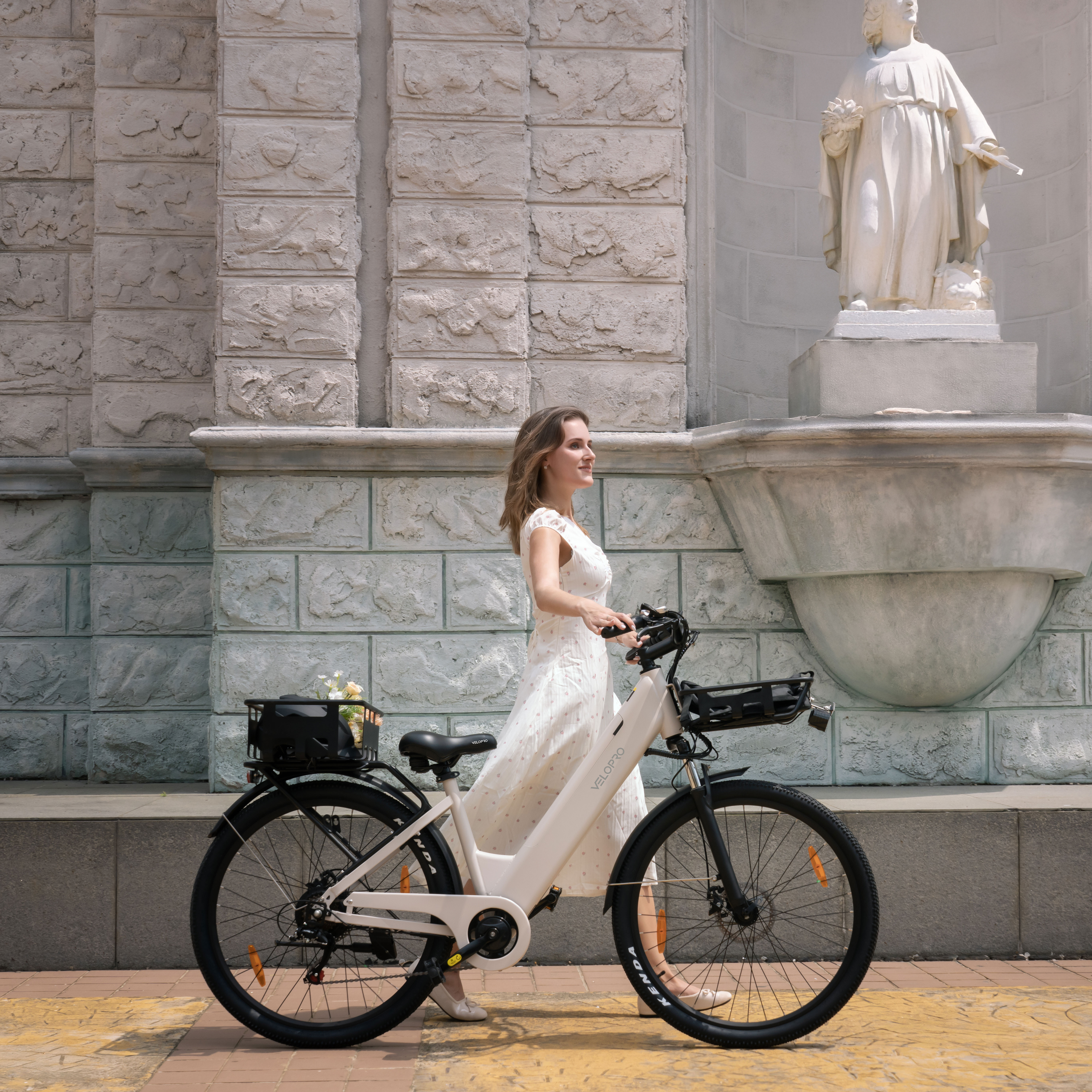 Una donna in abito bianco spinge una bicicletta elettrica bianca davanti a un muro di pietra e a una statua.