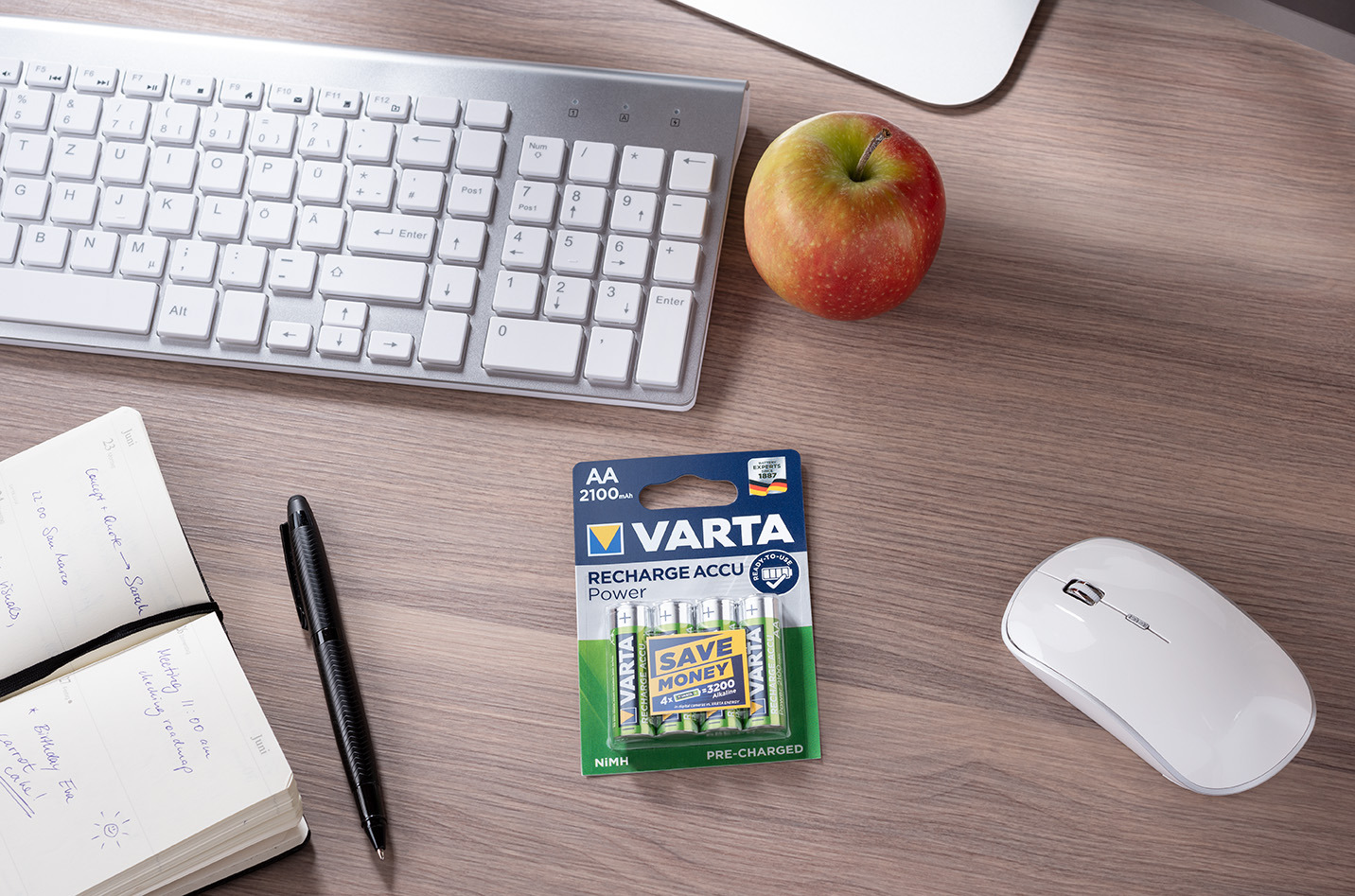 Escritorio de madera con teclado blanco, ratón, cuaderno, bolígrafo, una manzana y pilas Varta.