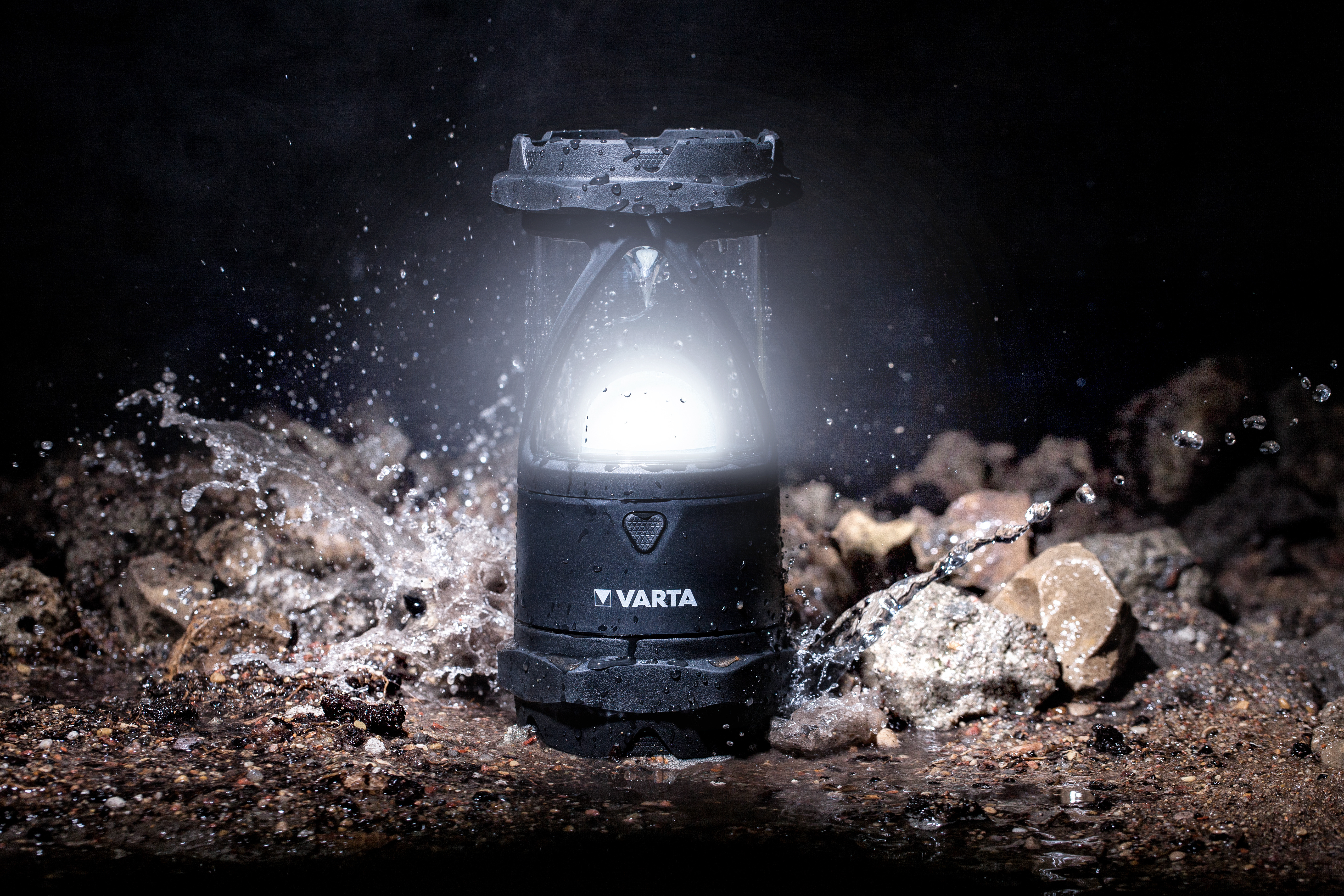 Eine schwarze Varta-Campinglaterne steht im Dunkeln auf nassem Boden, umgeben von spritzendem Wasser und Steinen.