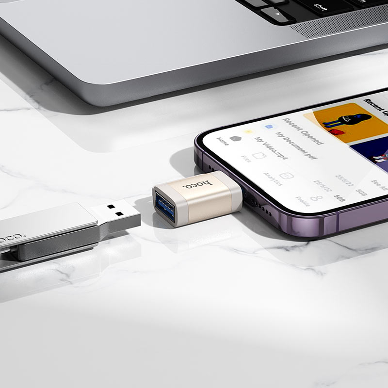 Ein Smartphone mit USB-Adapter und USB-Stick auf einem weißen Marmortisch, im Hintergrund ein Laptop.