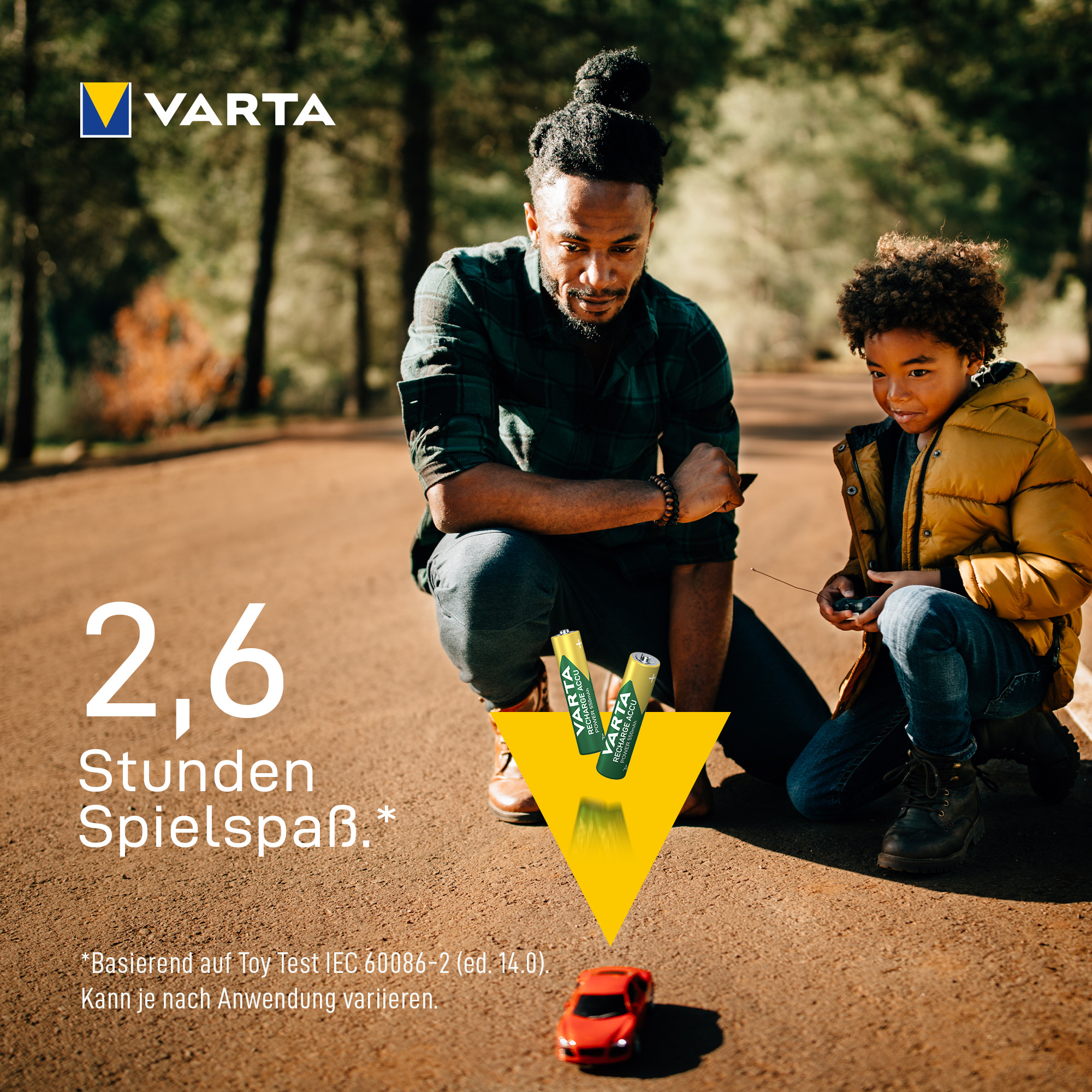 Ein Vater und sein Sohn spielen mit einem roten Spielzeugauto auf einem Waldweg, Varta-Werbung.