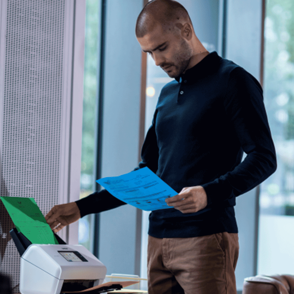 Ein Mann im dunklen Polohemd steht am Scanner, hält ein blaues Dokument und legt grünes Papier ein.