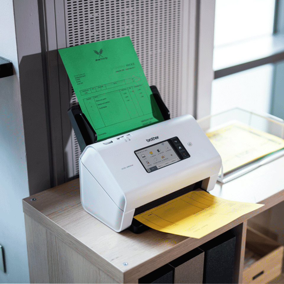 Ein weißer Brother-Scanner steht auf einem Holztisch, ein grünes Blatt ragt oben heraus und ein gelbes unten.