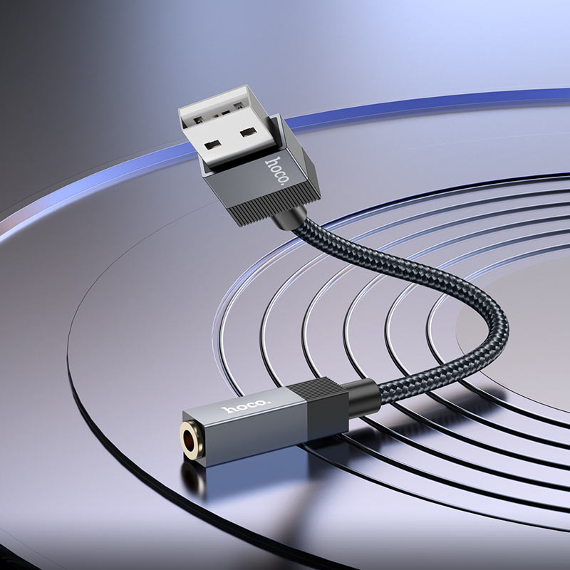 Un cavo USB-jack audio grigio con cordino intrecciato si trova su una superficie circolare riflettente.