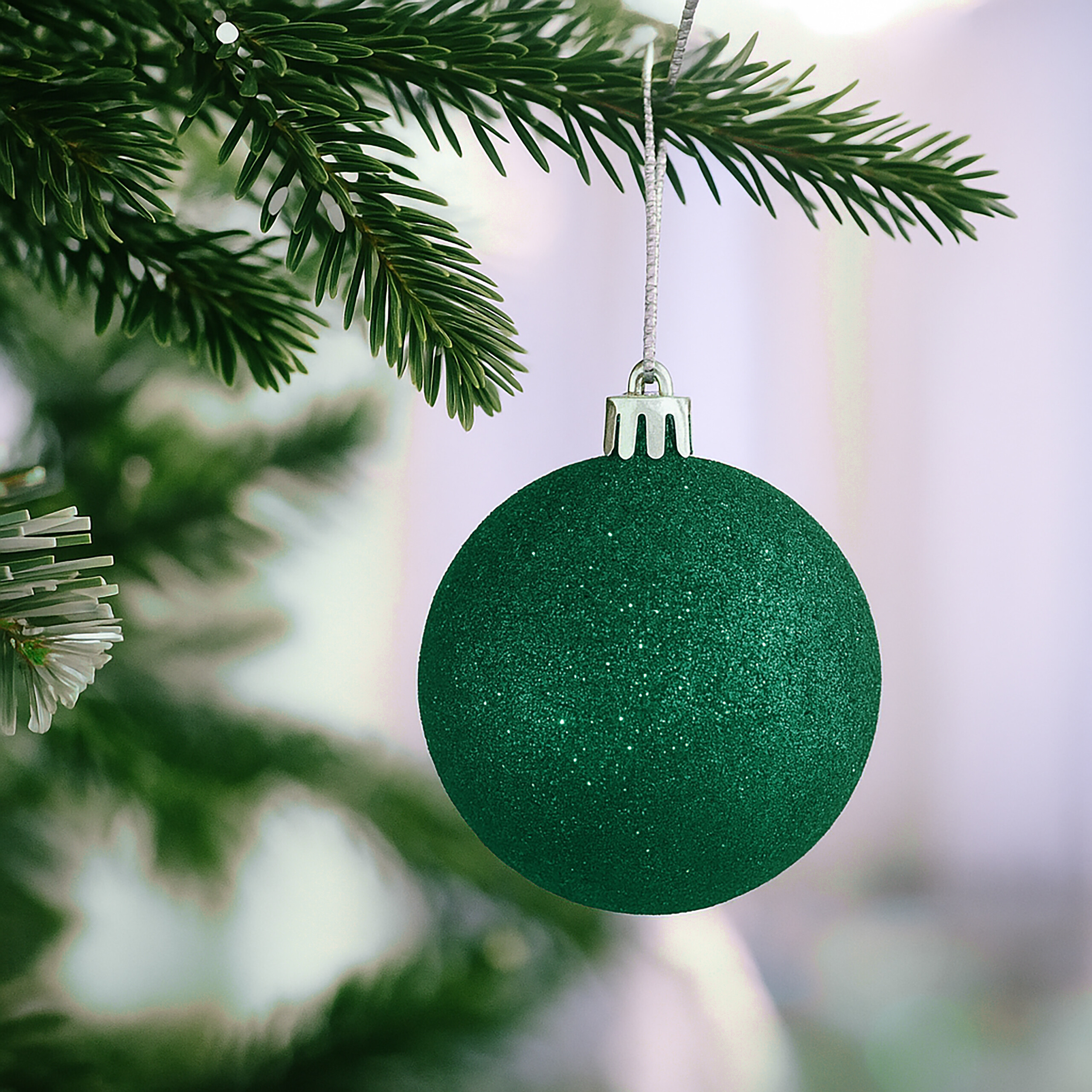 Eine glitzernde grüne Weihnachtskugel hängt an einem schneebedeckten Tannenzweig.