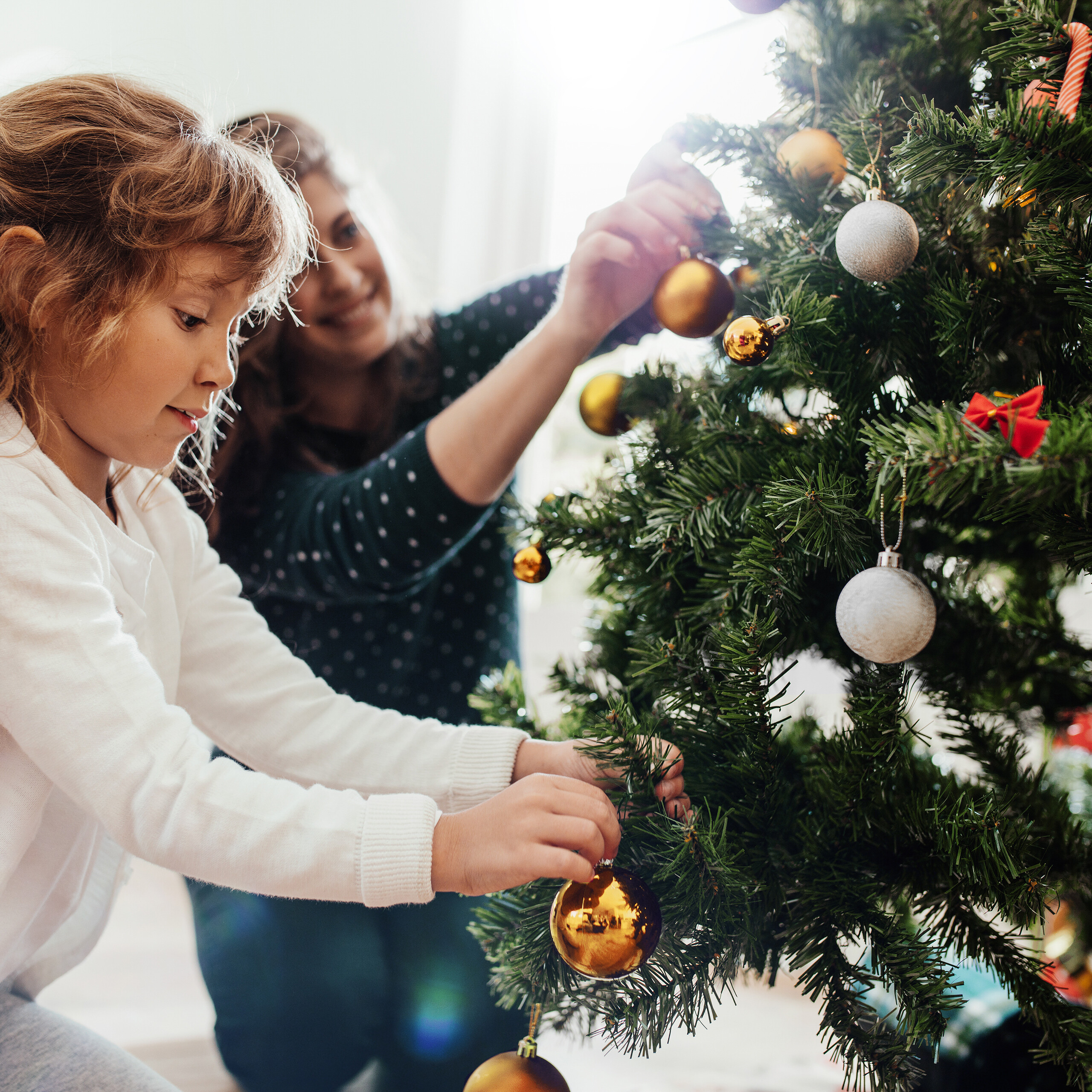Eine Mutter und ihre Tochter schmücken gemeinsam einen Weihnachtsbaum mit goldenen und silbernen Kugeln.