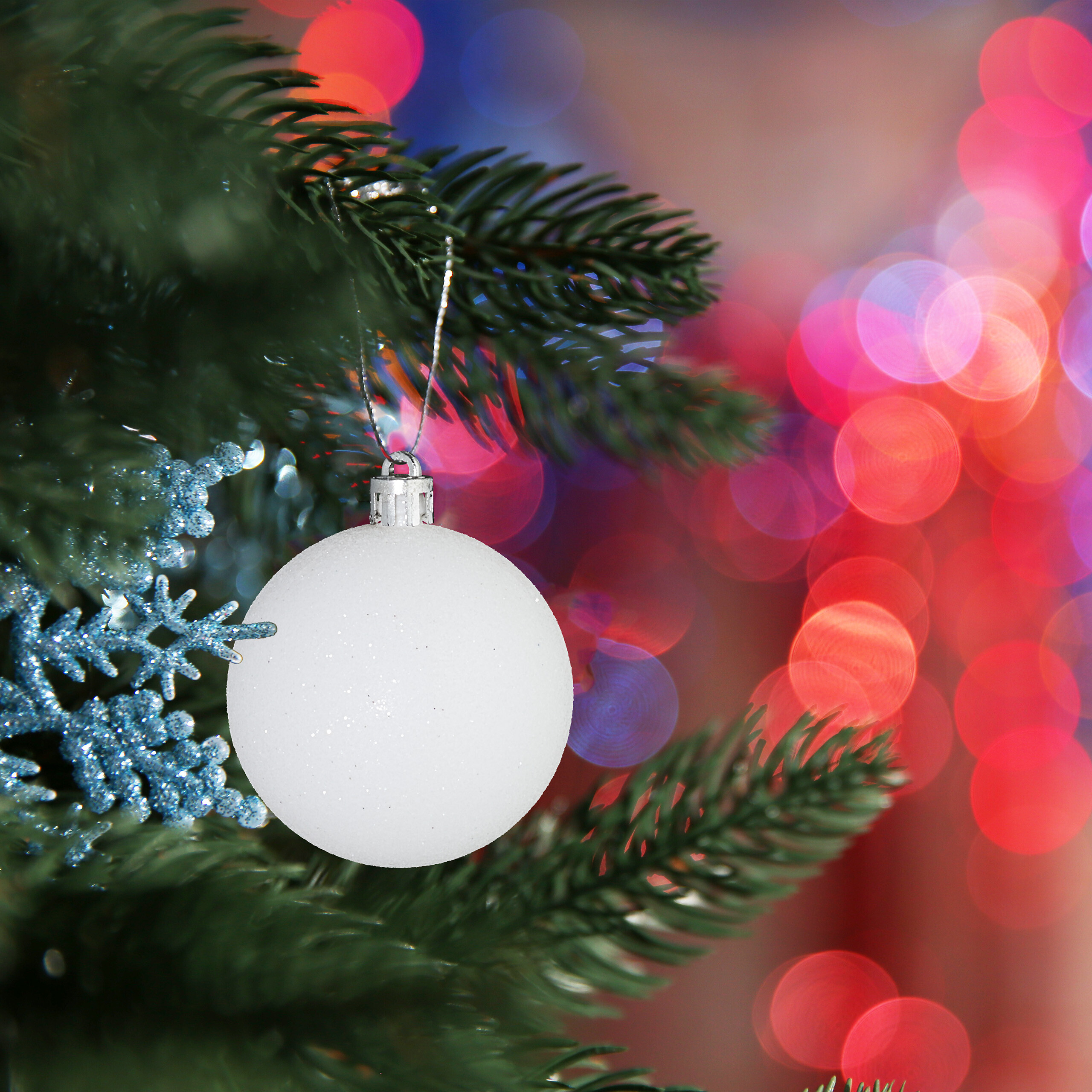 Eine weiße Weihnachtskugel hängt an einem Tannenast, mit blauen Schneeflocken und verschwommenem roten Hintergrund.