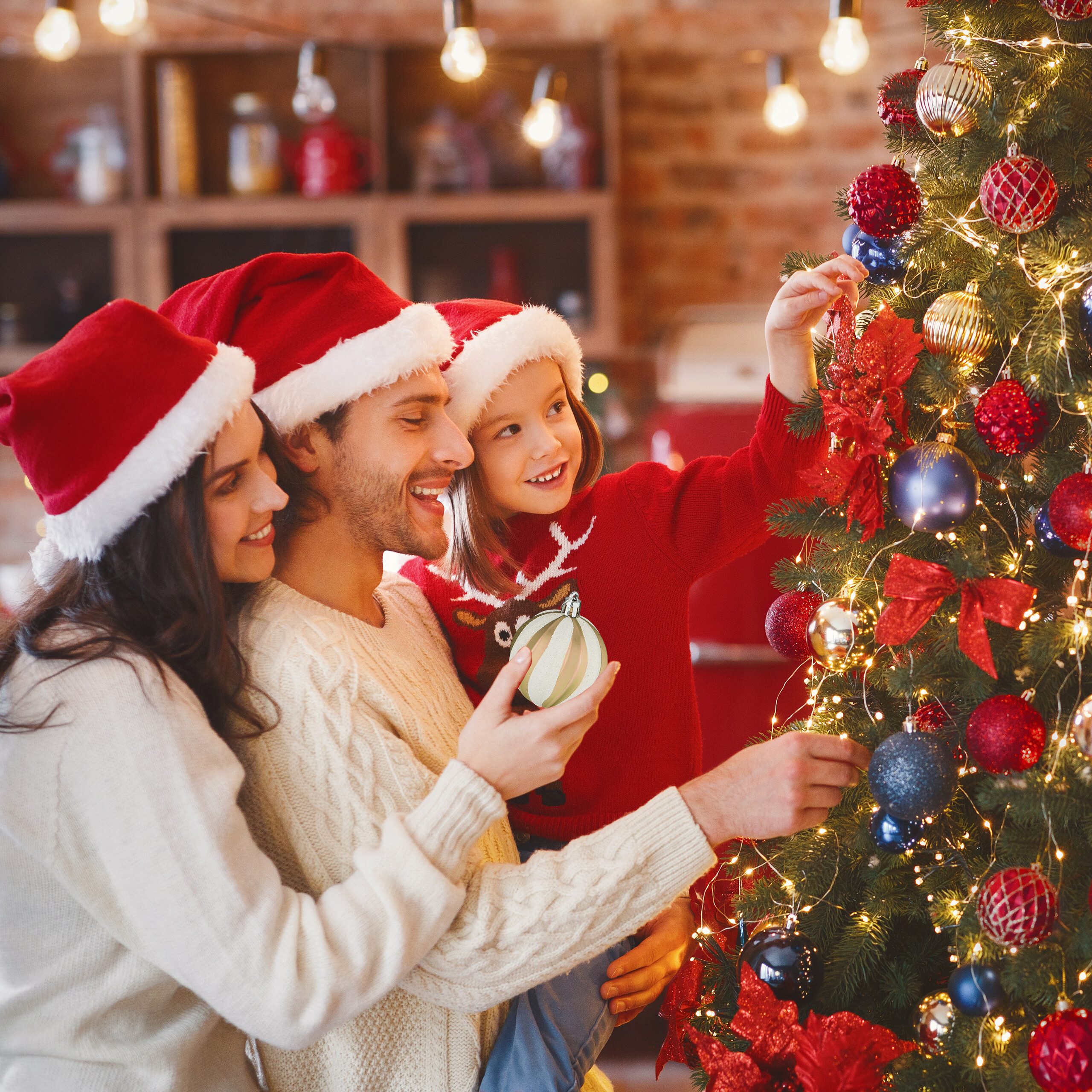 Eine Familie mit Weihnachtsmützen schmückt gemeinsam einen Weihnachtsbaum in einem gemütlichen Raum.