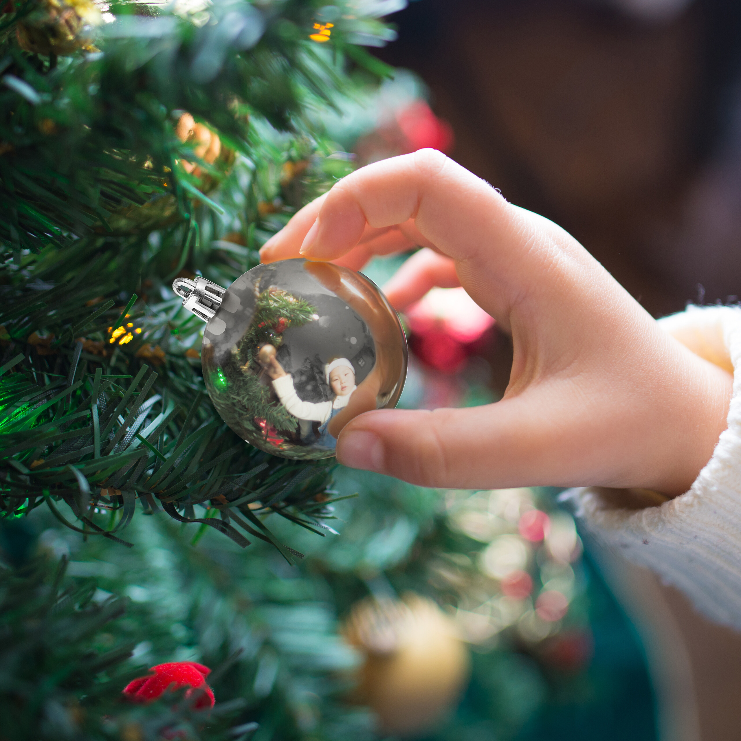 Die Hand eines Kindes hängt eine glänzende silberne Christbaumkugel an einen grünen Baum.