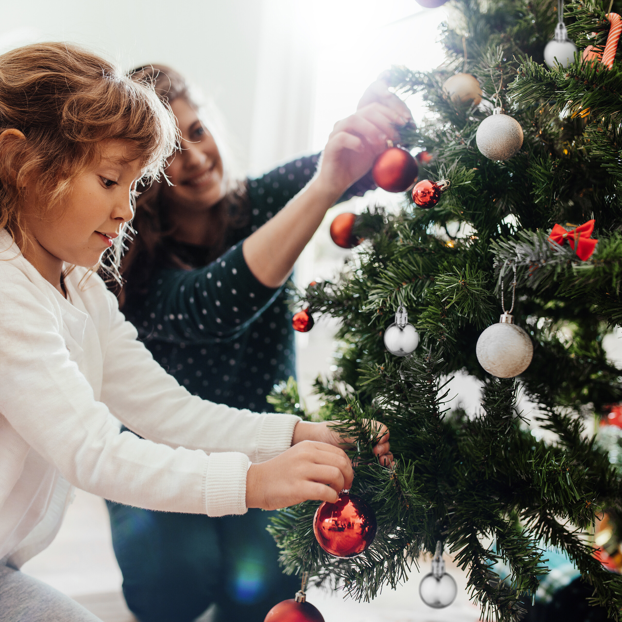 Ein Mädchen und eine Frau schmücken gemeinsam einen Weihnachtsbaum mit roten und silbernen Kugeln.