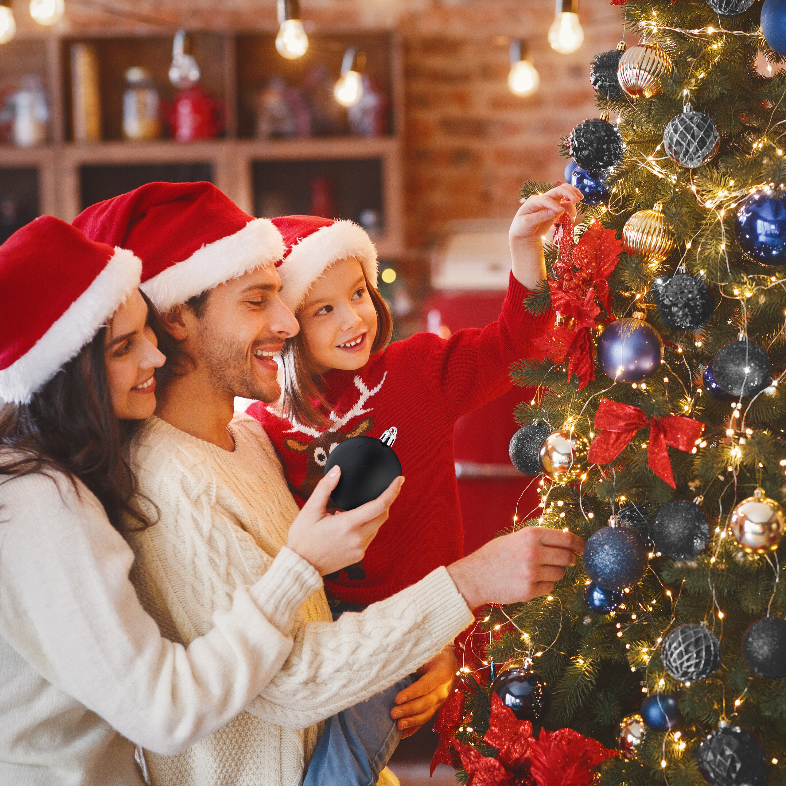 Eine Familie mit Weihnachtsmützen schmückt gemeinsam einen Weihnachtsbaum in einem gemütlichen Raum.