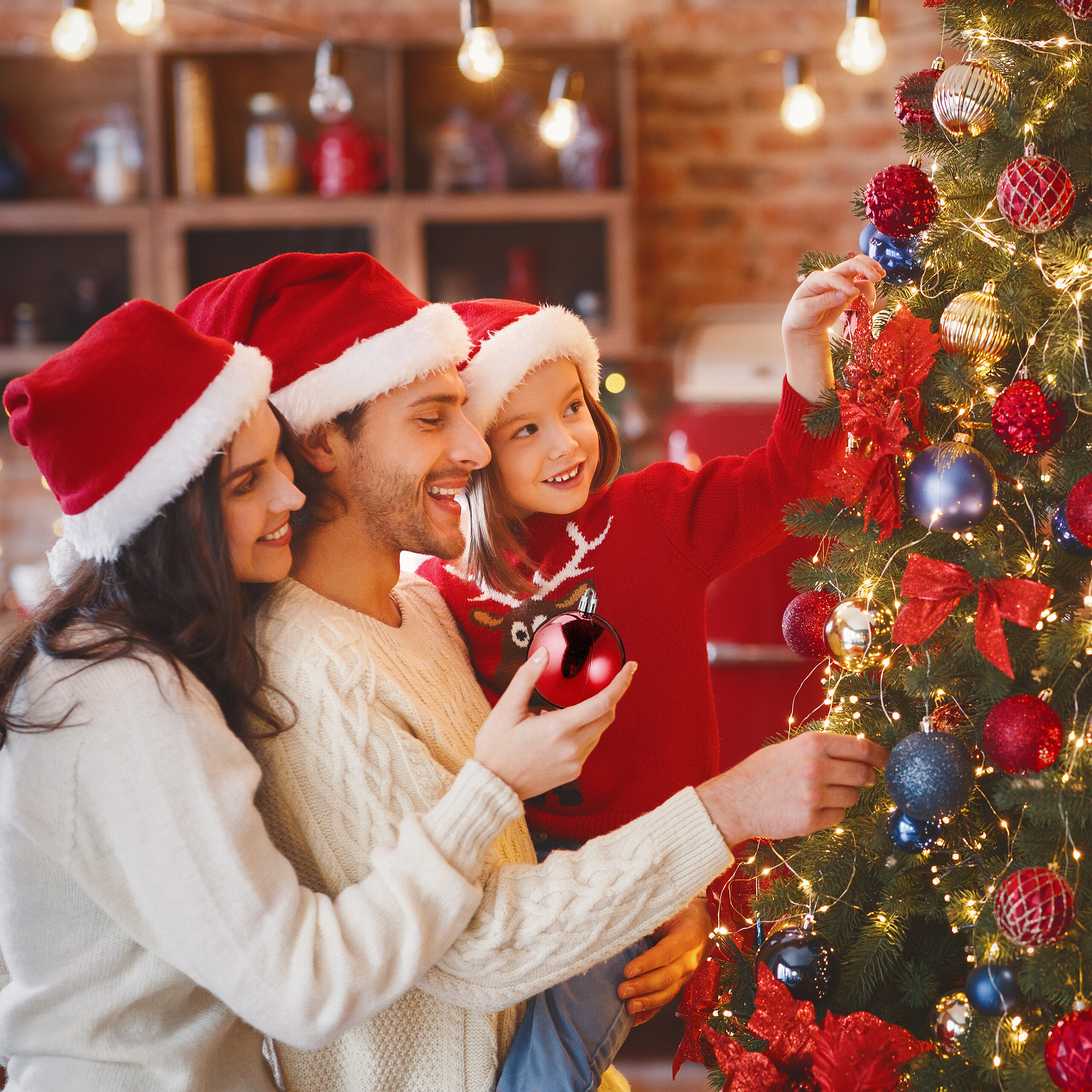 Eine Familie mit Weihnachtsmützen dekoriert gemeinsam einen Weihnachtsbaum in einem gemütlichen Raum.