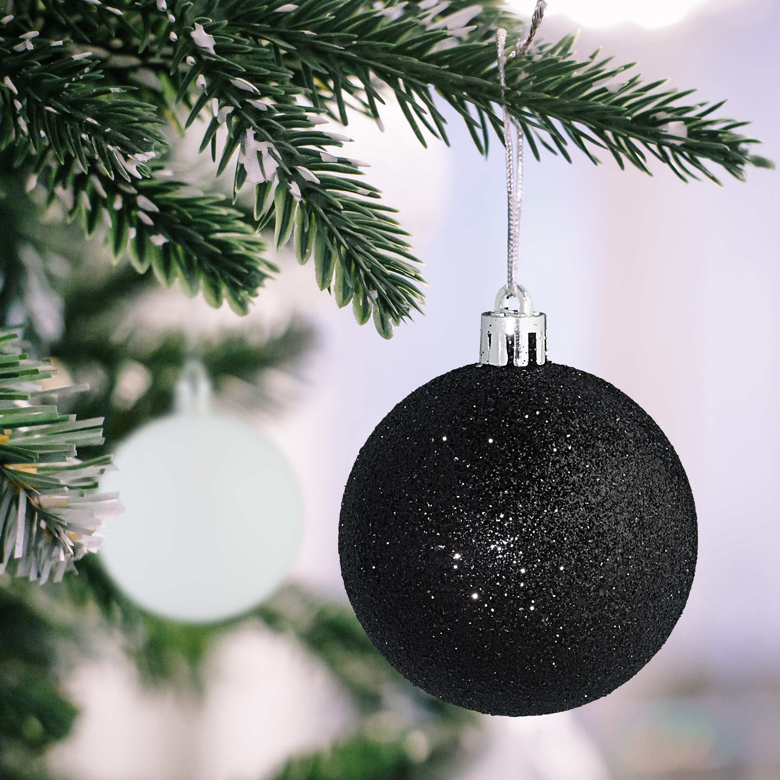 Eine glitzernde schwarze Christbaumkugel hängt an einem schneebedeckten Tannenzweig, im Hintergrund ein verschwommener Ball.