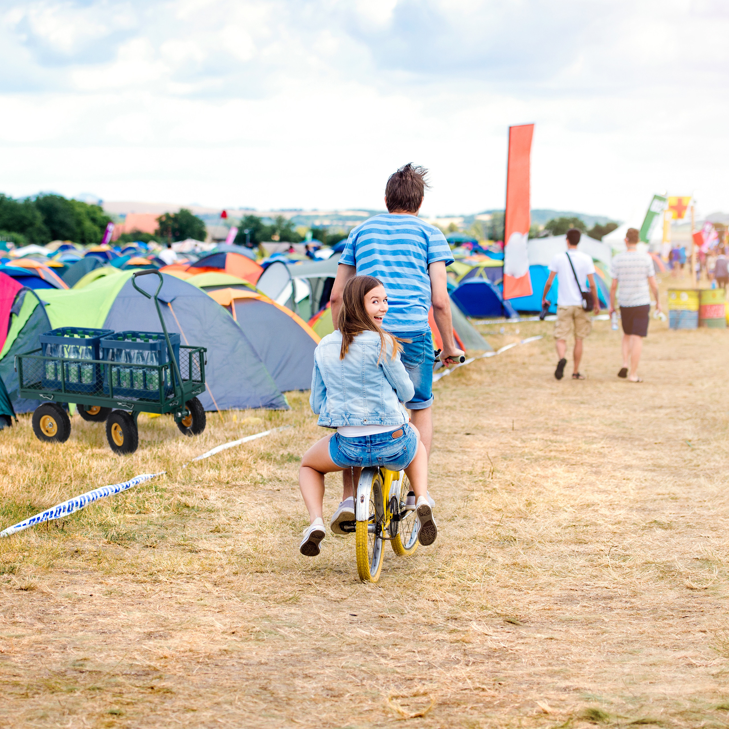 Ein Paar fährt auf einem Fahrrad durch ein Festivalgelände mit vielen bunten Zelten im Hintergrund.