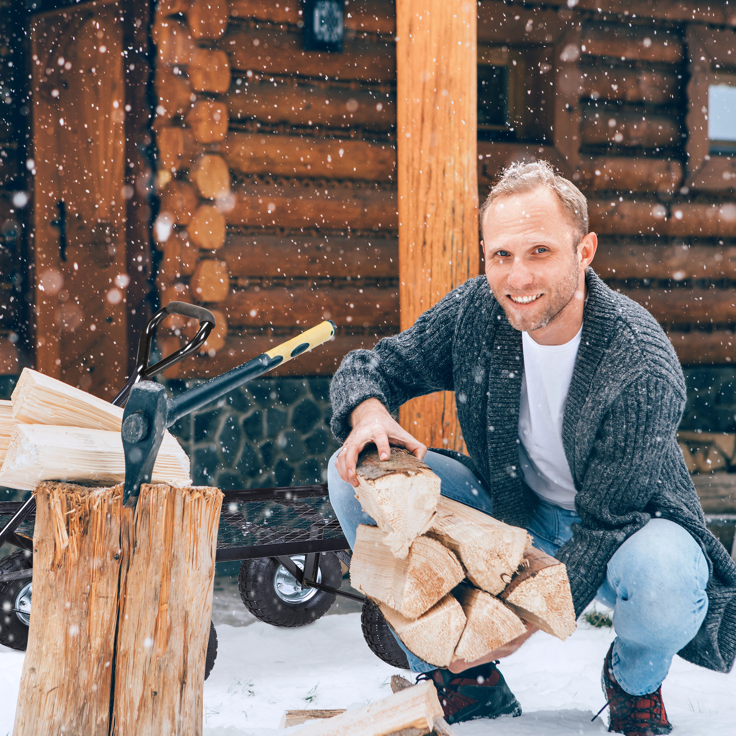 Ein Mann im grauen Cardigan kniet im Schnee und hält Kaminholz vor einer Holzhütte.
