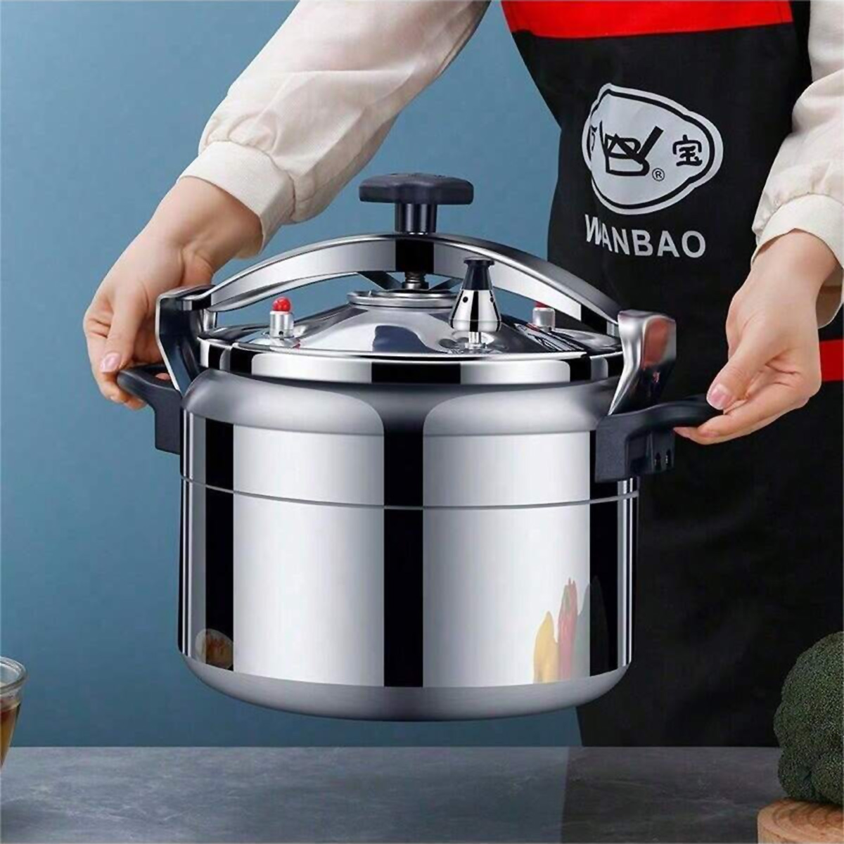 Persona sosteniendo una olla a presión de acero inoxidable brillante con mango negro sobre fondo azul.