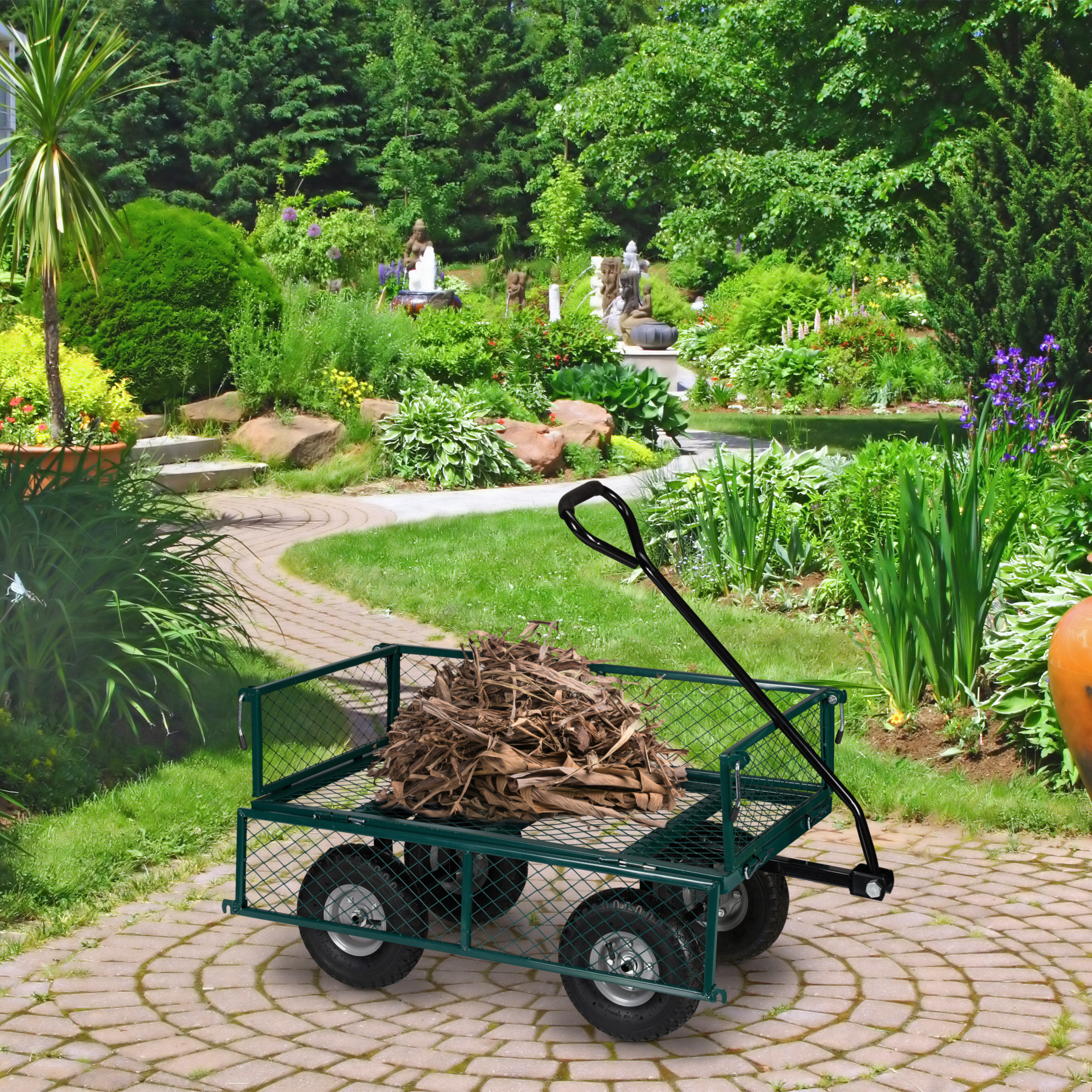 Ein grüner Gartenwagen voller trockener Blätter steht auf einem Steinweg vor einer üppigen Gartenlandschaft.