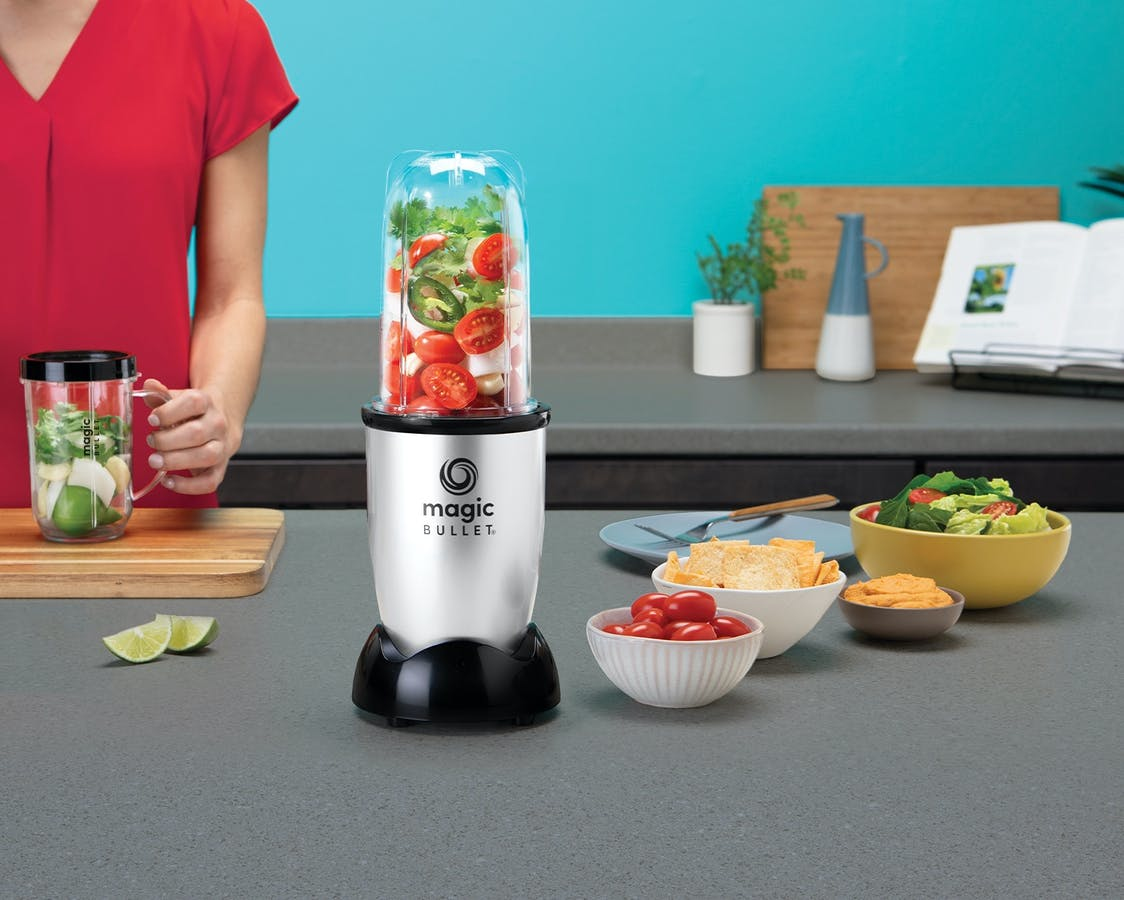 A Magic Bullet blender filled with vegetables sits on a kitchen counter with snacks.