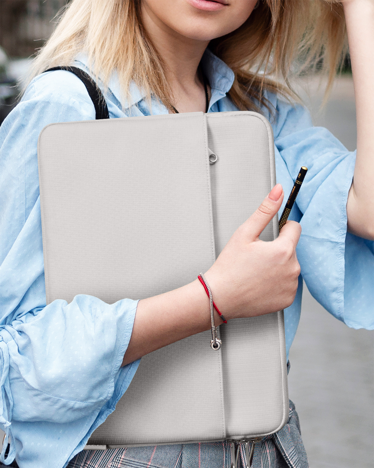 Eine blonde Frau im blauen Hemd hält eine graue Laptoptasche und einen Bleistift.