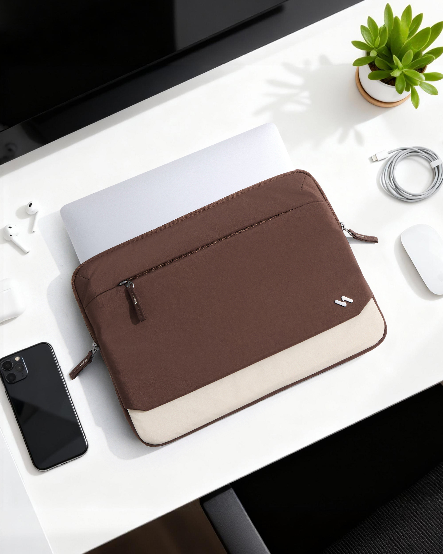 Eine braune Laptoptasche mit einem hellen Streifen liegt auf einem weißen Schreibtisch neben Handy, Maus und Pflanze.