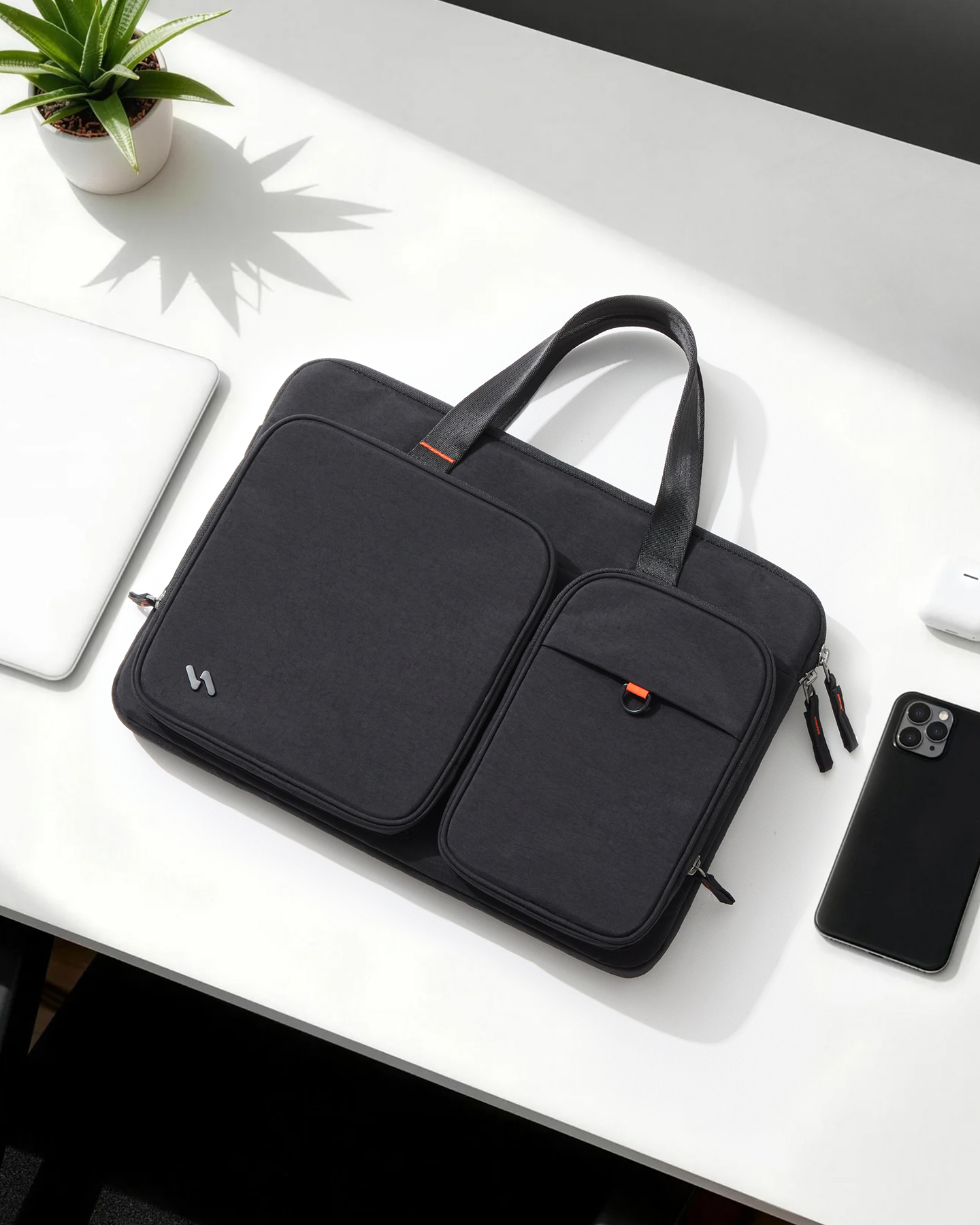 Eine schwarze Laptoptasche mit Vordertaschen und Griffen liegt auf einem weißen Schreibtisch neben einem Smartphone und einer Pflanze.