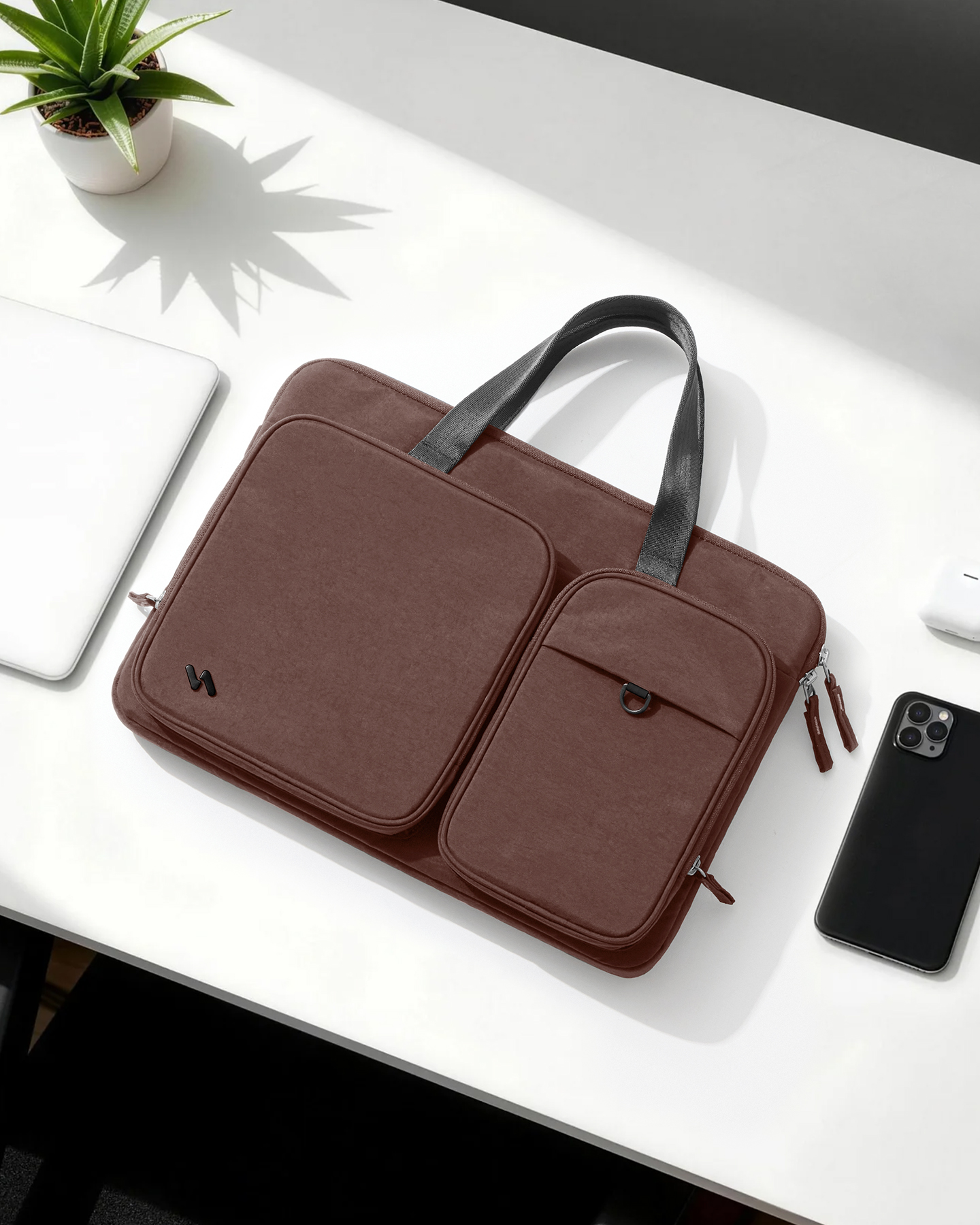 Eine braune Laptoptasche mit Vordertaschen auf einem weißen Schreibtisch mit Telefon und Topfpflanze.