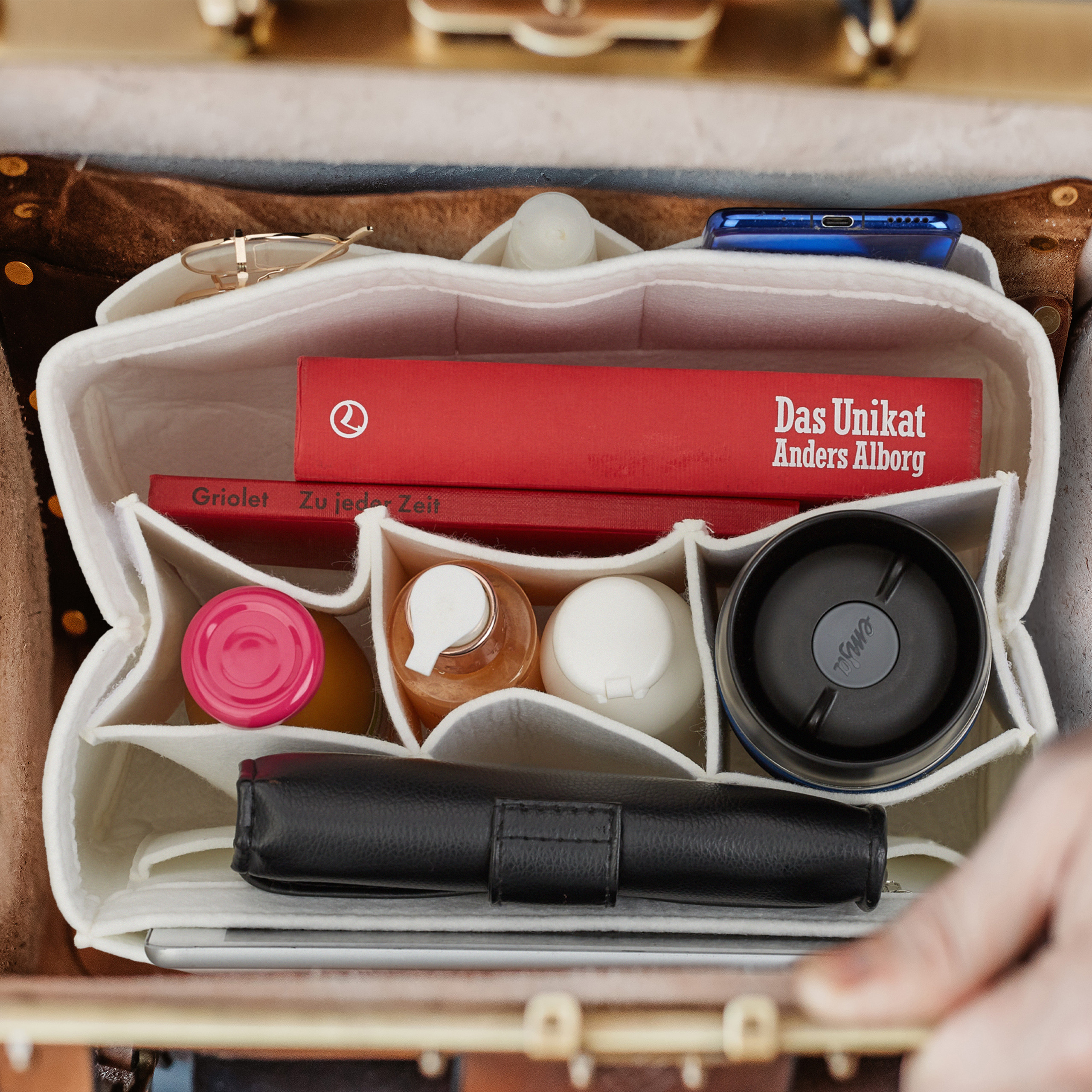Eine organisierte braune Ledertasche mit weißem Filz-Organizer, roten Büchern, Flaschen, Becher und schwarzer Geldbörse.