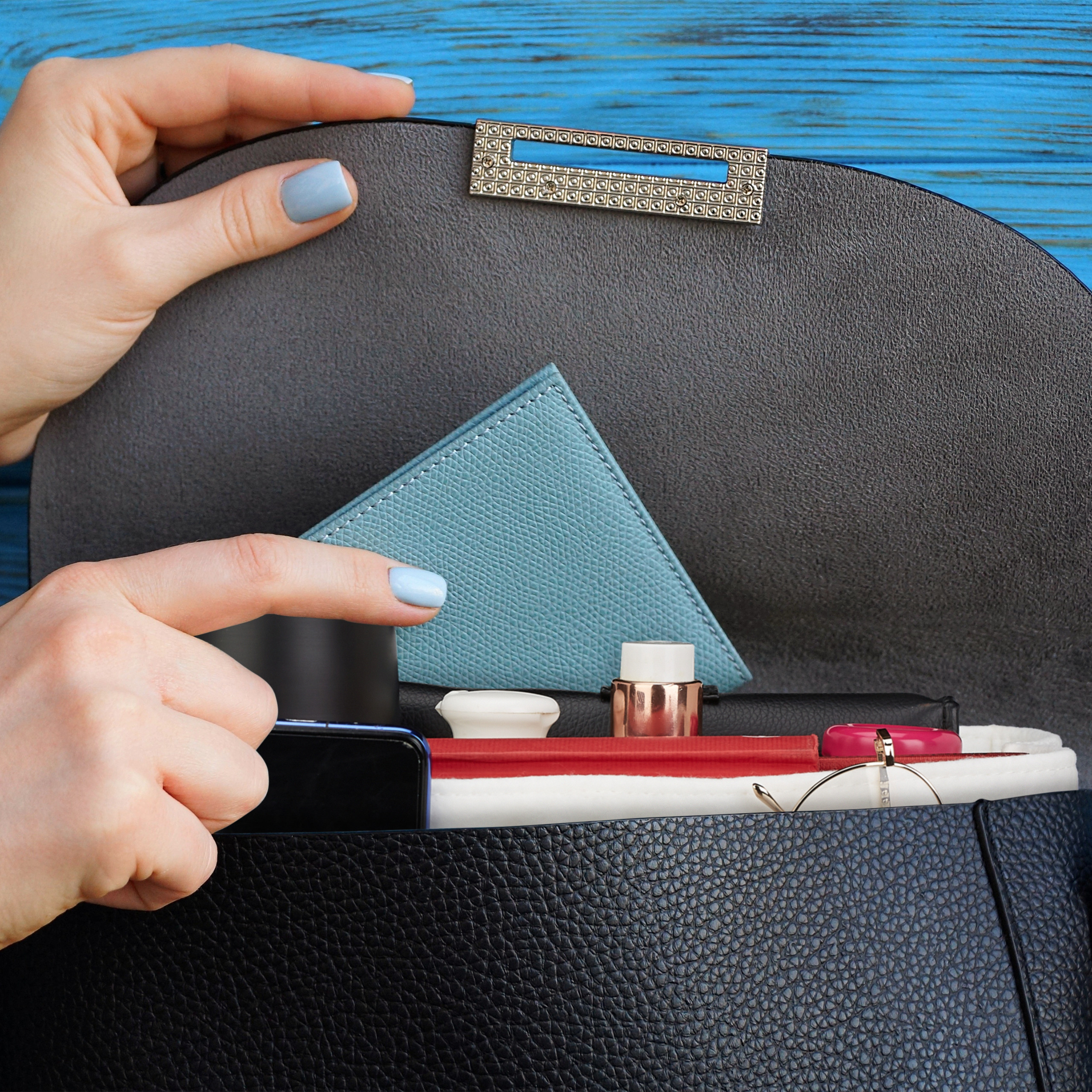 Eine Frau öffnet eine schwarze Lederhandtasche mit einer blauen Geldbörse, Kosmetik und einem Handy vor blauer Holzwand.