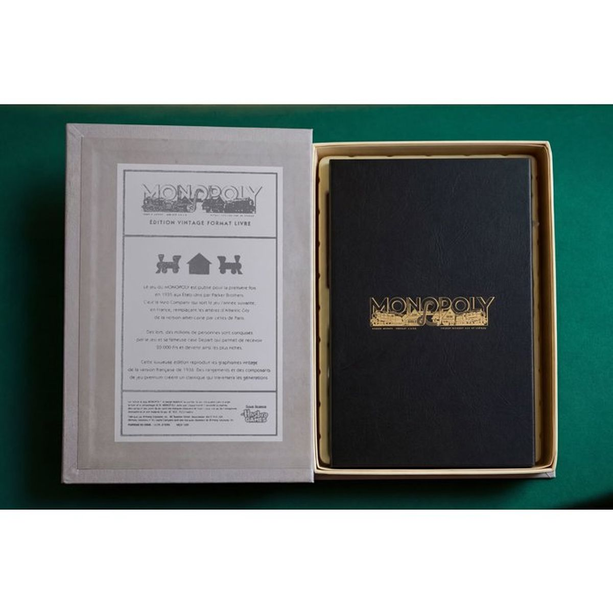 Eine geöffnete Vintage-Monopoly-Spielbox im Buchformat zeigt einen schwarzen Einband mit goldenem Logo auf einem grünen Tisch.