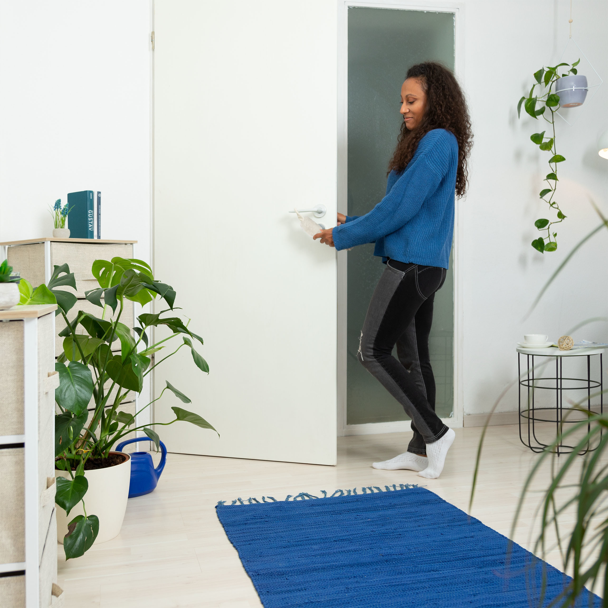 Eine Frau in blauem Pullover öffnet eine weiße Tür in einem hellen Raum mit Pflanzen.