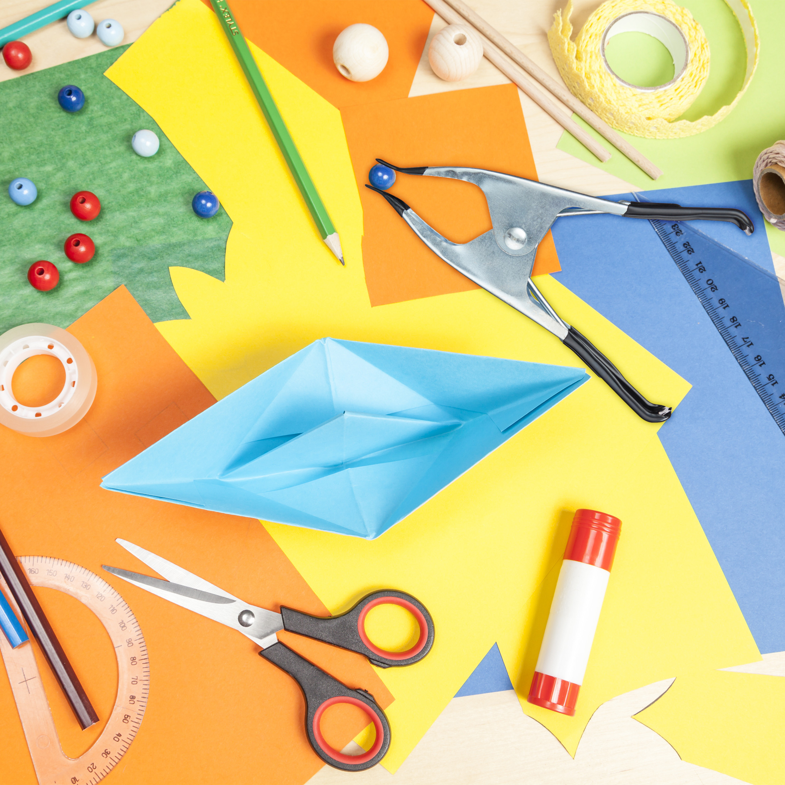 Bunte Bastelmaterialien mit Papierschiff, Schere, Klebestift und Perlen auf einem Tisch.