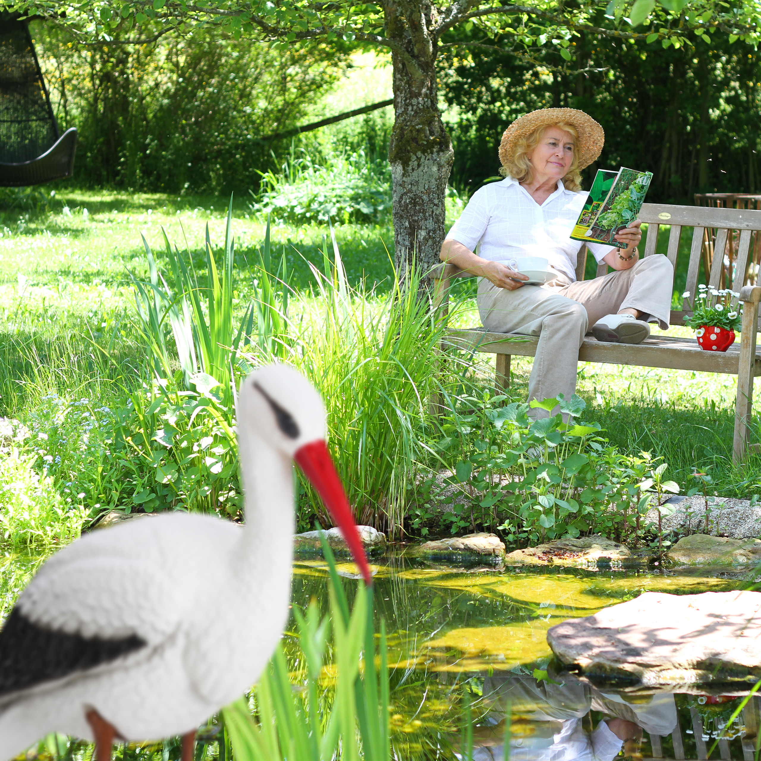 Eine Frau liest ein Magazin auf einer Holzbank im Garten, während im Vordergrund eine künstliche Storchfigur am Teich steht.