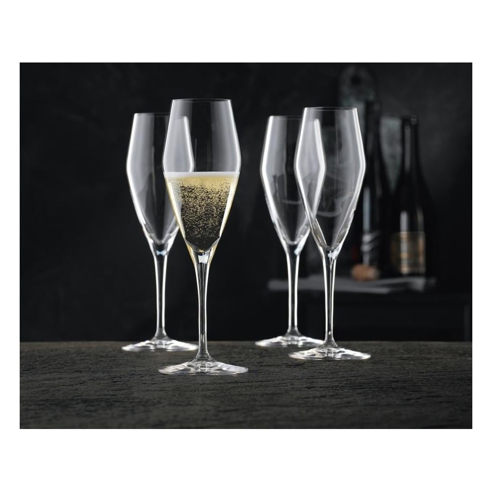 Vier Sektgläser auf dunklem Tisch, eines gefüllt mit prickelndem Sekt und Flaschen im Hintergrund.