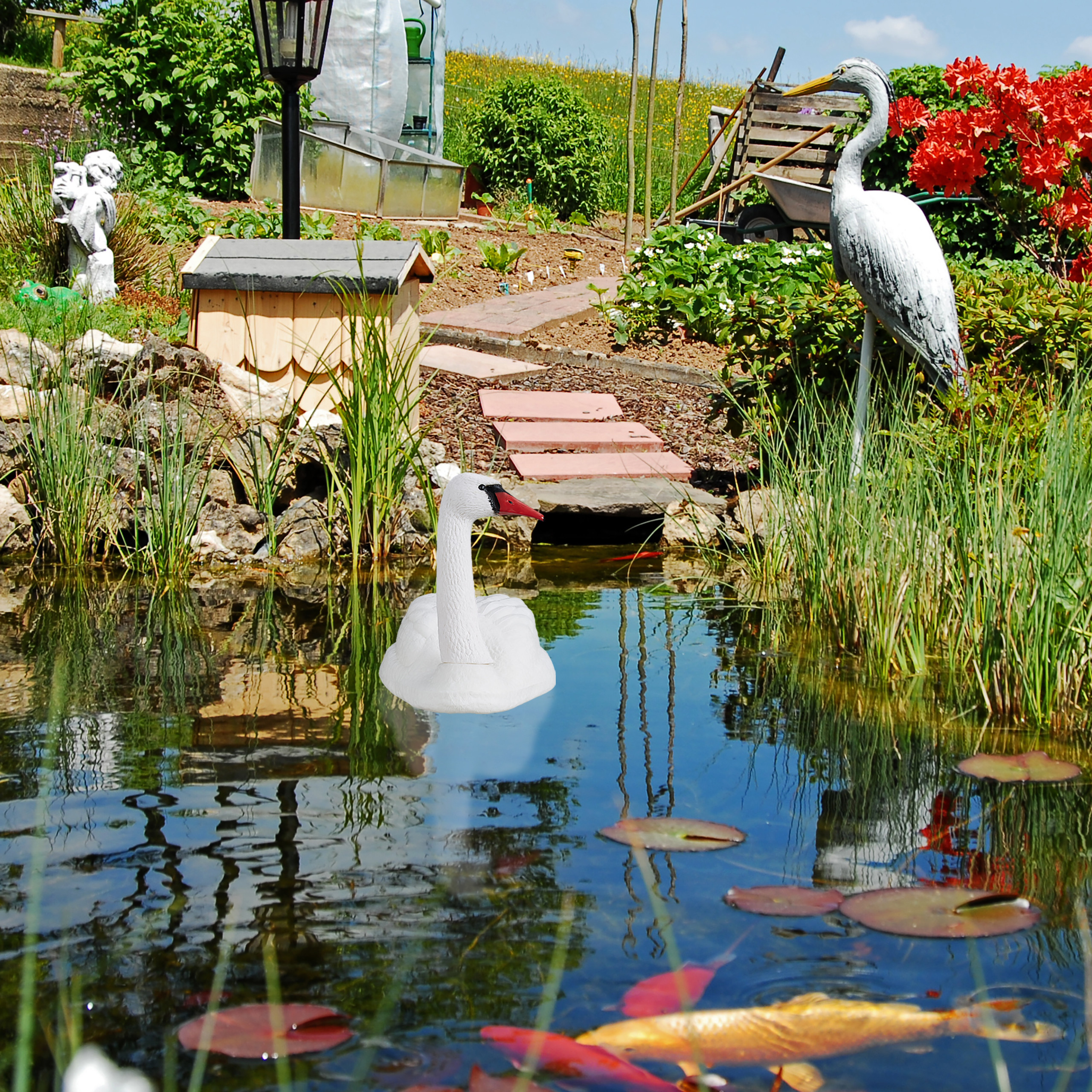 Ein Gartenteich mit einer weißen Schwanenattrappe, einer Reiherstatue, Seerosen und bunten Koifischen im Wasser.