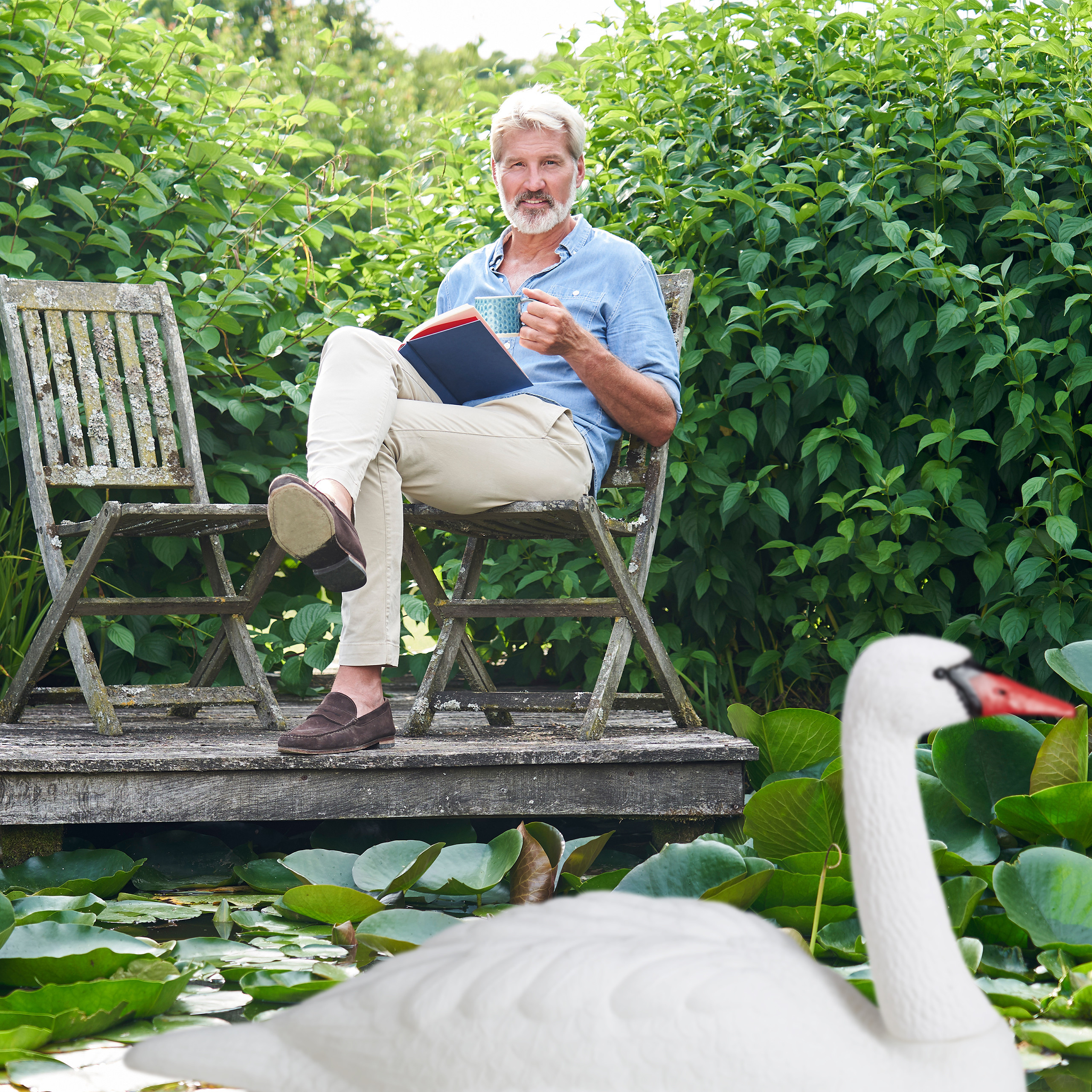 Ein älterer Mann liest ein Buch und hält eine Tasse auf einer Holzbank am Teich mit einem Spielzeugschwan.