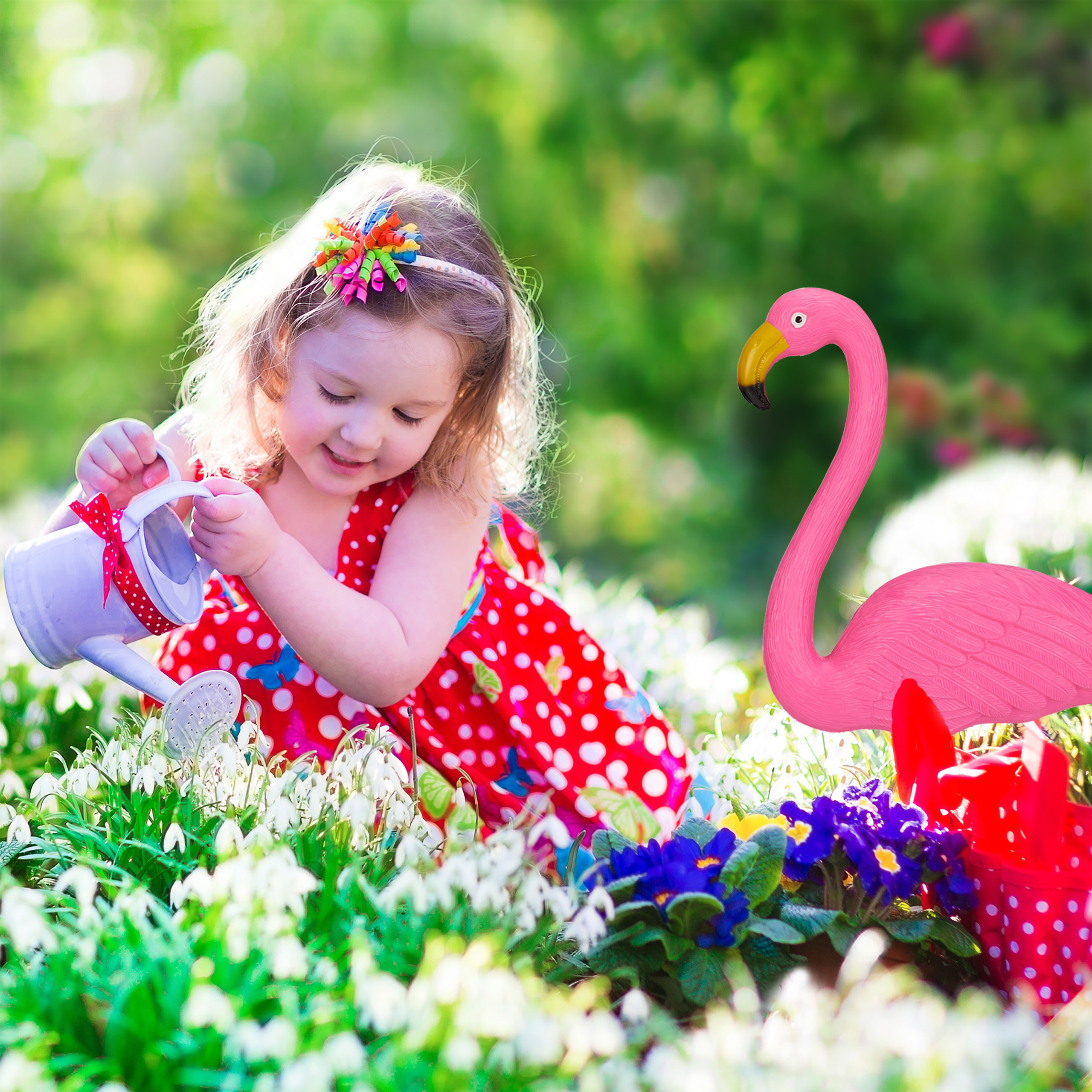 Kleines Mädchen in rotem Polka-Dot-Kleid gießt Blumen im Garten neben einer rosa Flamingo-Figur.